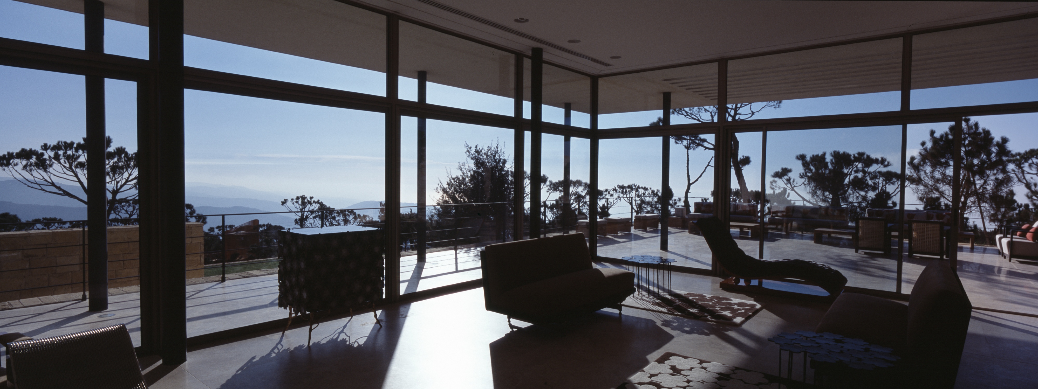 Living room of F House in Dahr el Sawan, floor-to-ceiling glazing framing pine trees and hazy hilltop views at dusk