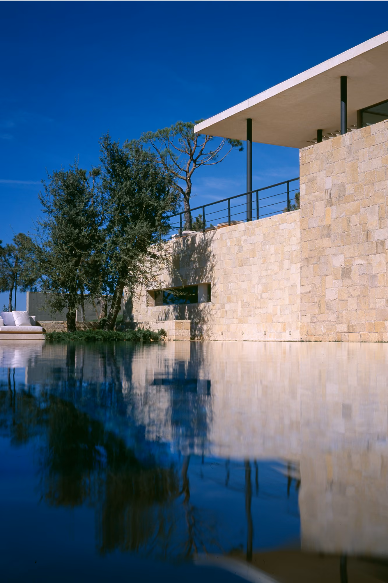 Reflecting pool beside stone-clad walls of F House in Dahr el Sawan, with a cantilevered flat roof and pine trees under a clear blue sky