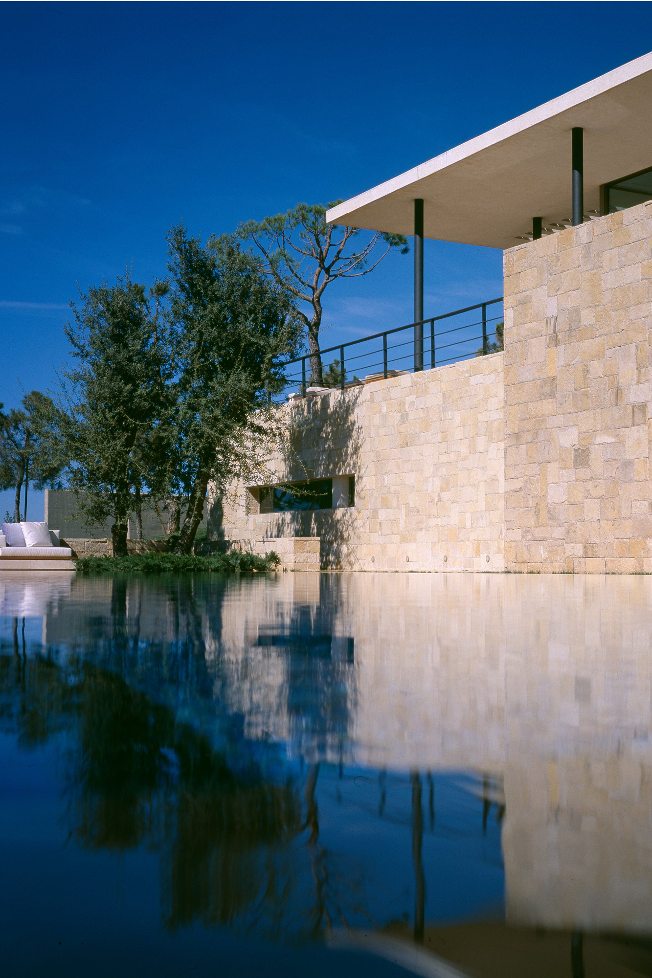 Reflecting pool beside stone-clad walls of F House in Dahr el Sawan, with a cantilevered flat roof and pine trees under a clear blue sky