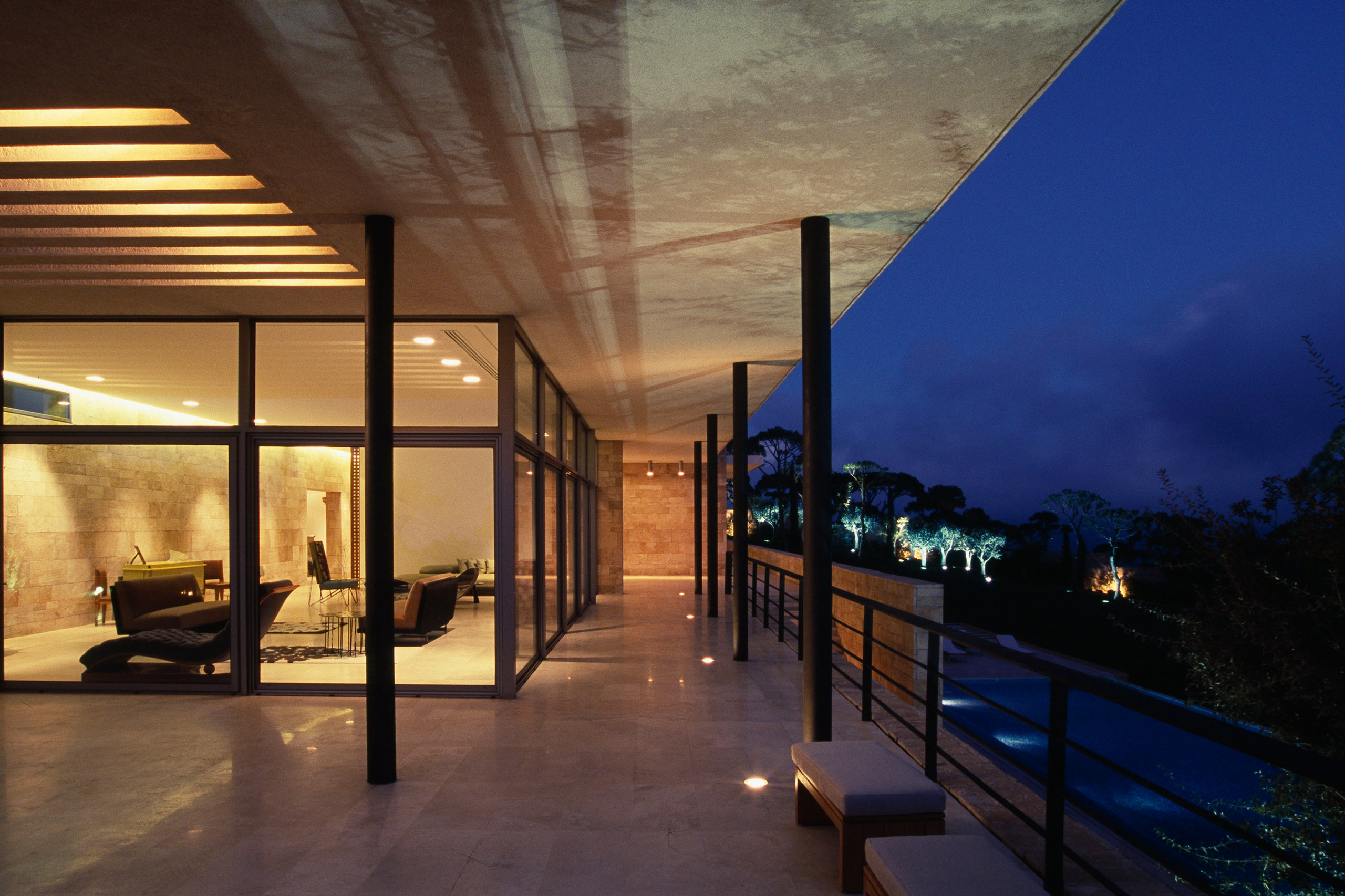 Illuminated covered terrace of F House in Dahr el Sawan at dusk, slender columns lining a walkway beside a pool and floodlit trees on the hillside