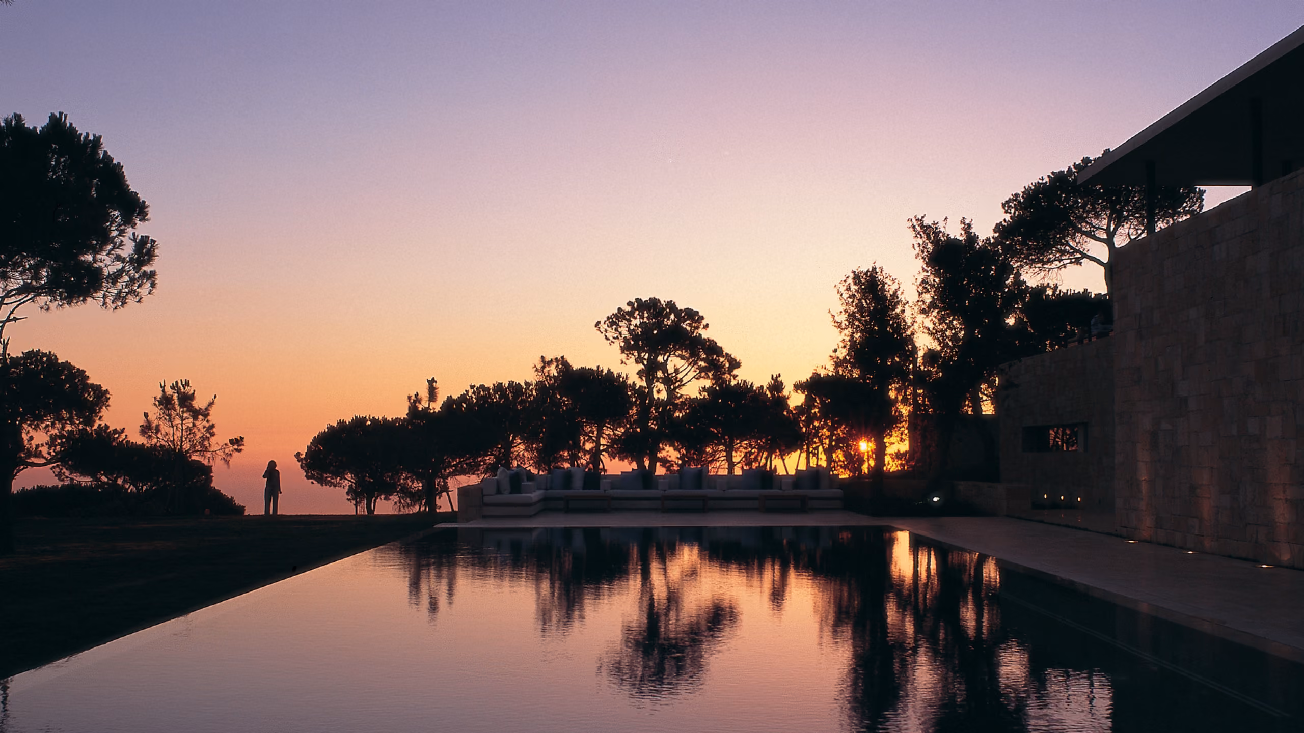 Twilight view of F House in Dahr el Sawan, reflecting pool bordered by stone walls and pine trees silhouetted against an orange and purple sunset sky