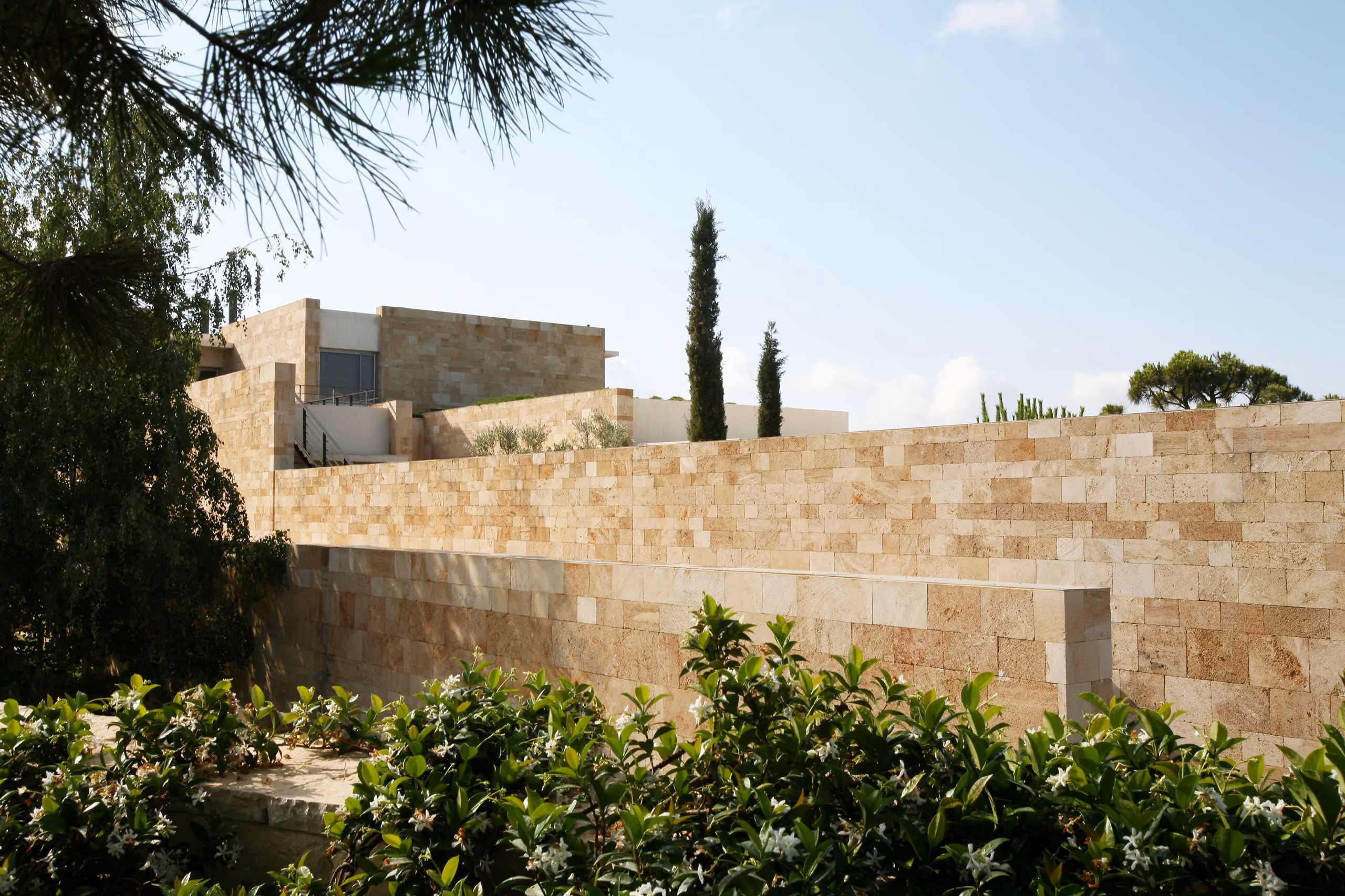 Local sandstone retaining walls of F House in Dahr el Sawan frame the stone-clad upper level, with cypress trees and flowering shrubs surrounding the hillside residence