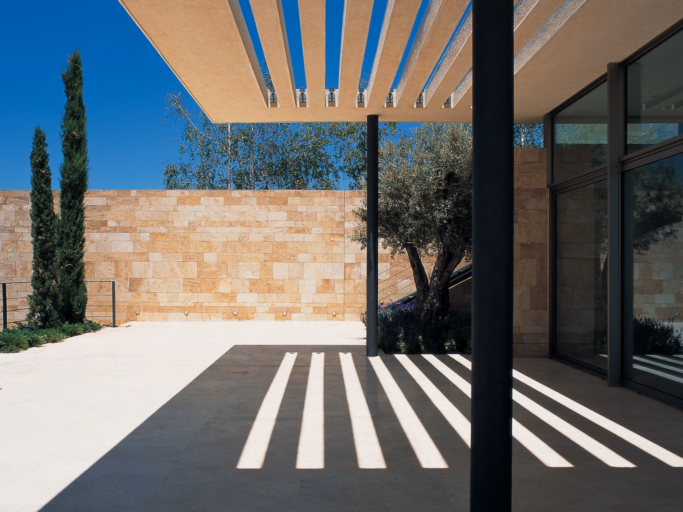 Terrace at F House in Dahr el Sawan, slatted canopy casting linear shadows over stone paving beside a sandstone wall and cypress trees