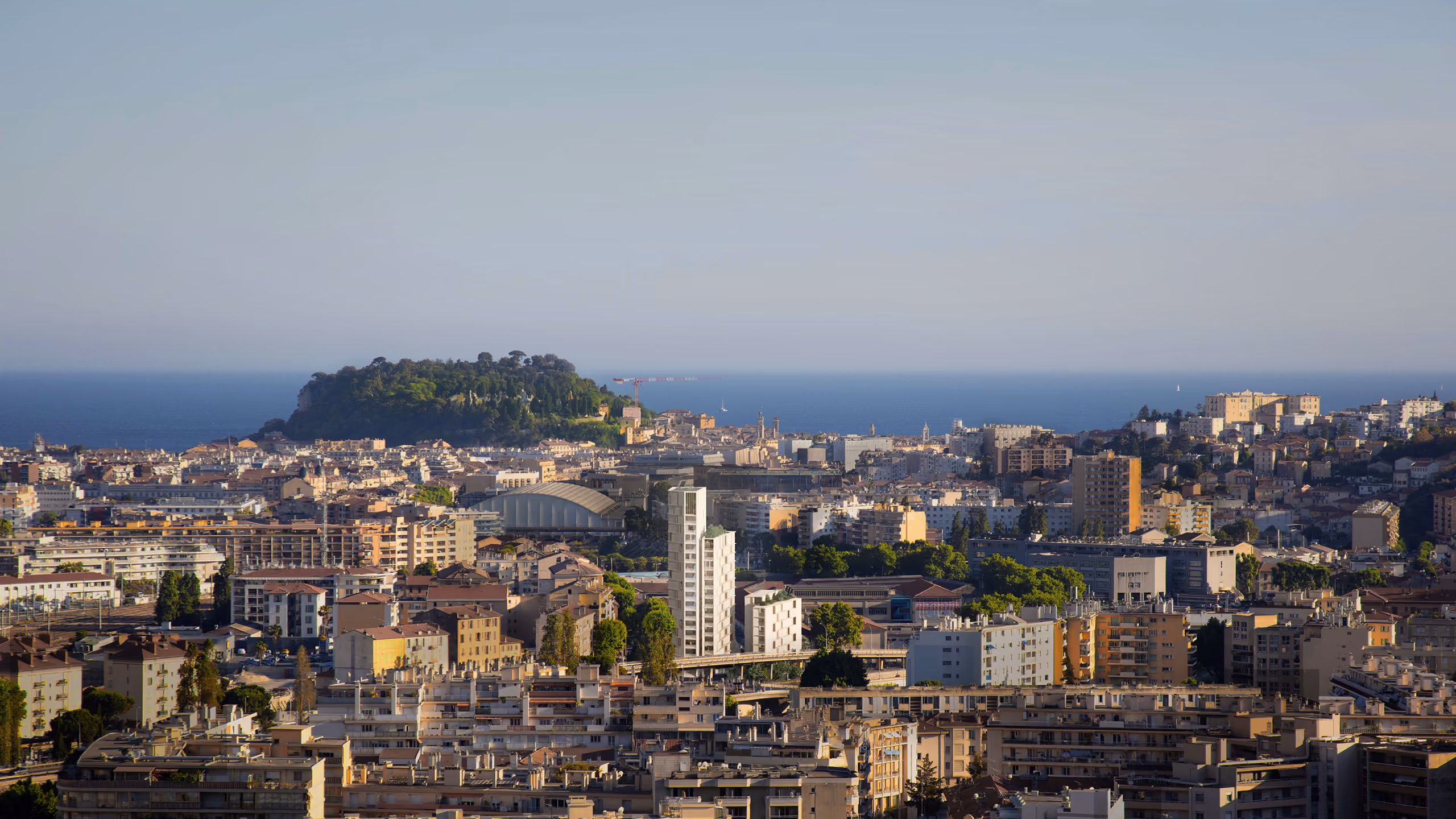 Aerial view of Tour de Nice, a slender residential tower rising above the dense rooftops of Nice, with a wooded headland and the Mediterranean Sea beyond