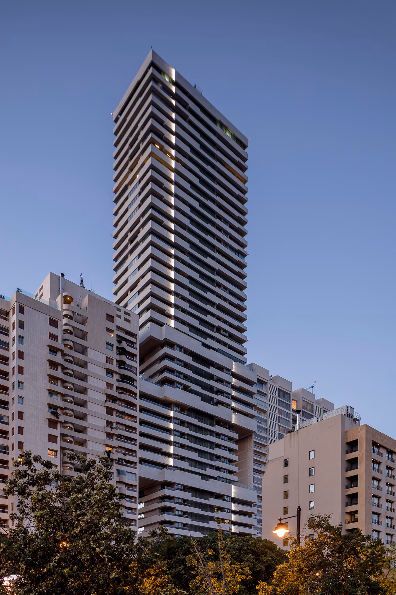Skygate tower in Beirut at dusk, its stacked cantilevered volumes lit with vertical light strips, rising above surrounding residential high-rises