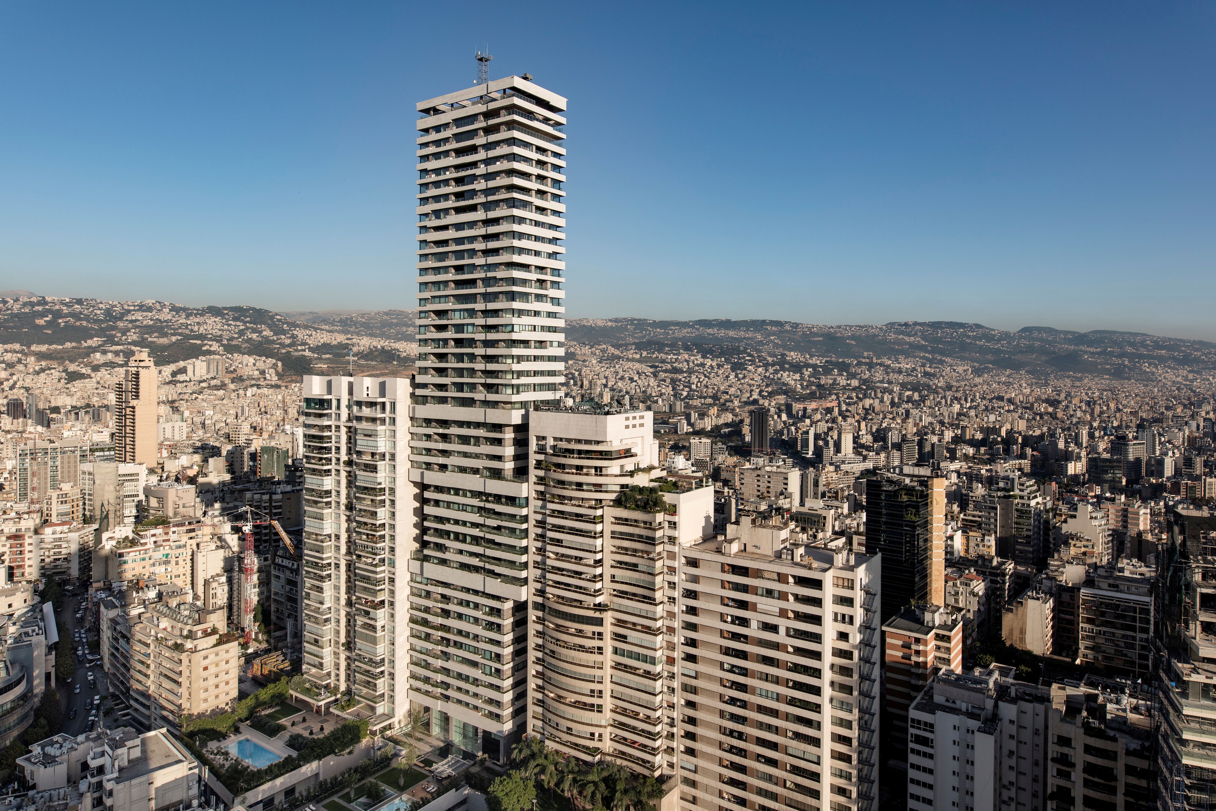 Skygate's stacked cantilevered volumes towering over Beirut's dense cityscape, with hills and haze visible in the distance