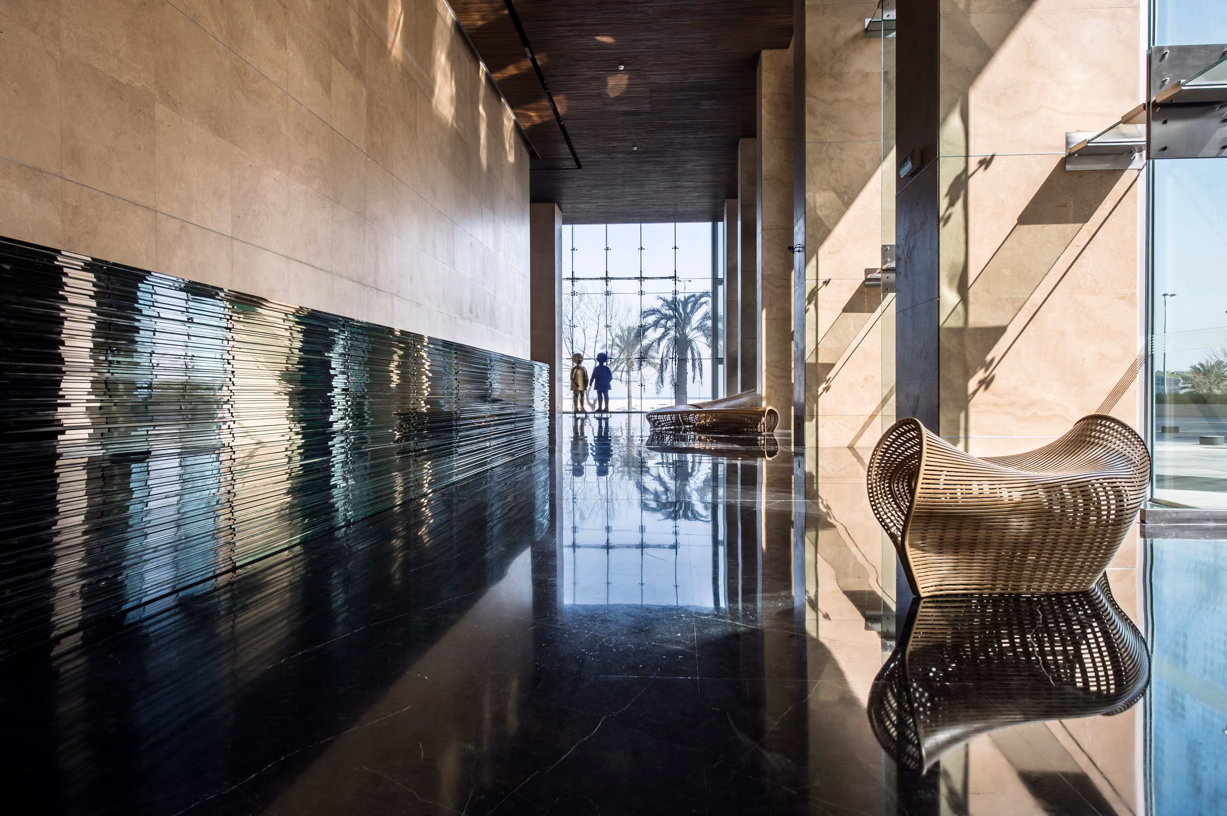 Lobby interior of Platinum Tower in Beirut, reflective dark stone floor, ribbed glass wall panel, woven sculptural benches and figures silhouetted against a palm-lined view
