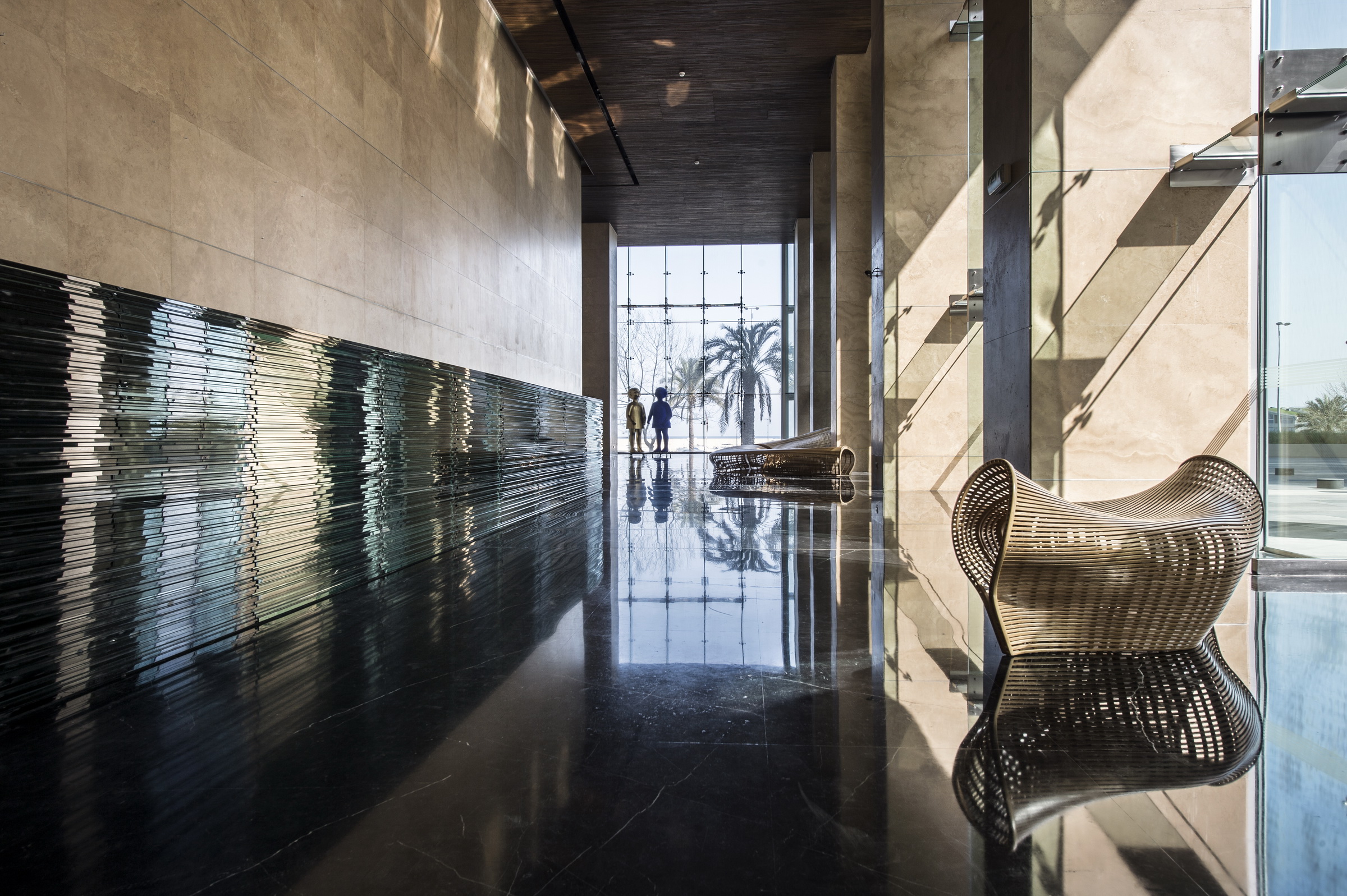 Lobby interior of Platinum Tower in Beirut, reflective dark stone floor, ribbed glass wall panel, woven sculptural benches and figures silhouetted against a palm-lined view
