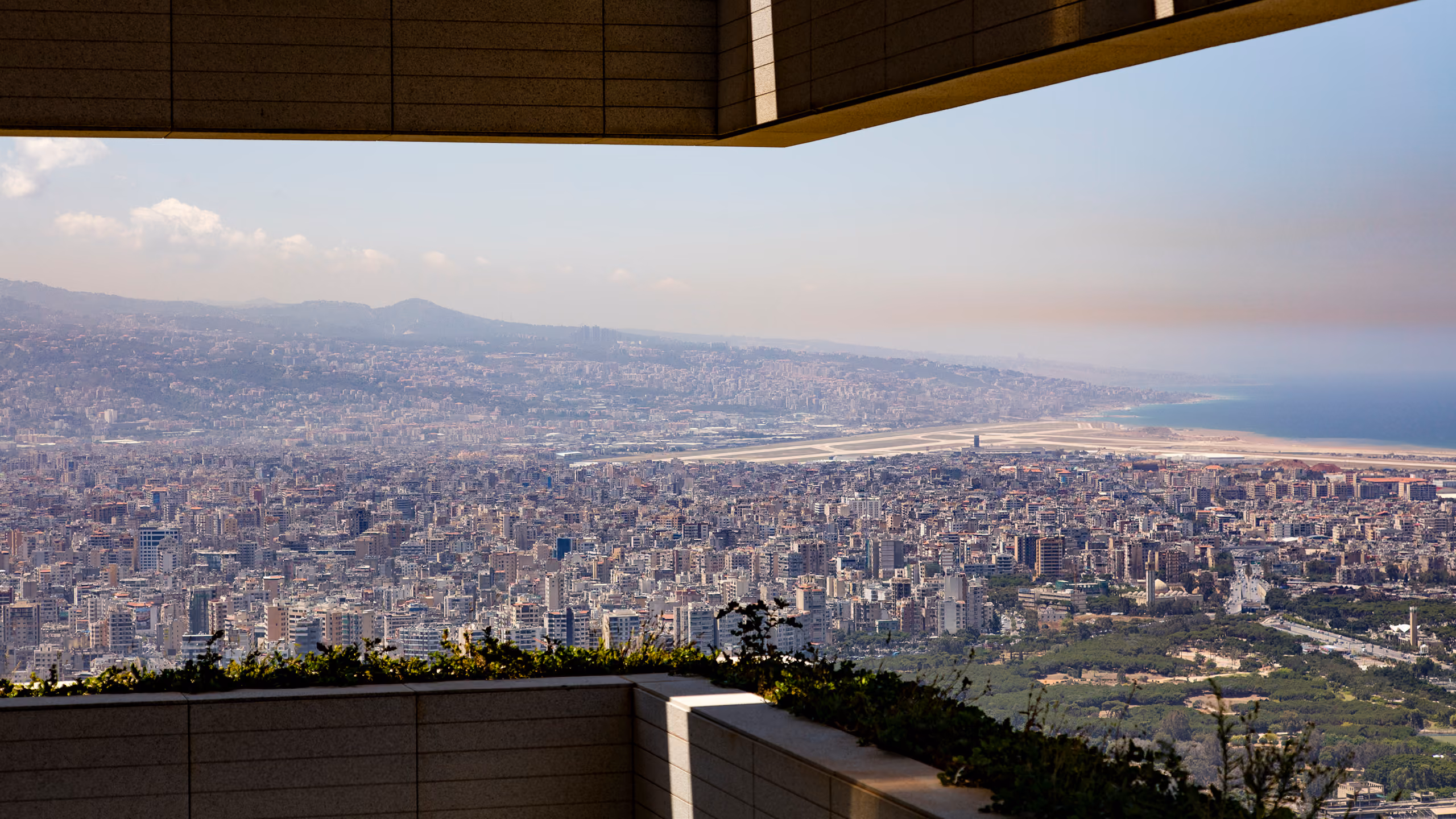 Sweeping hilltop view from a Skygate terrace in Beirut over dense cityscape toward the coastline and airport runway in hazy daylight