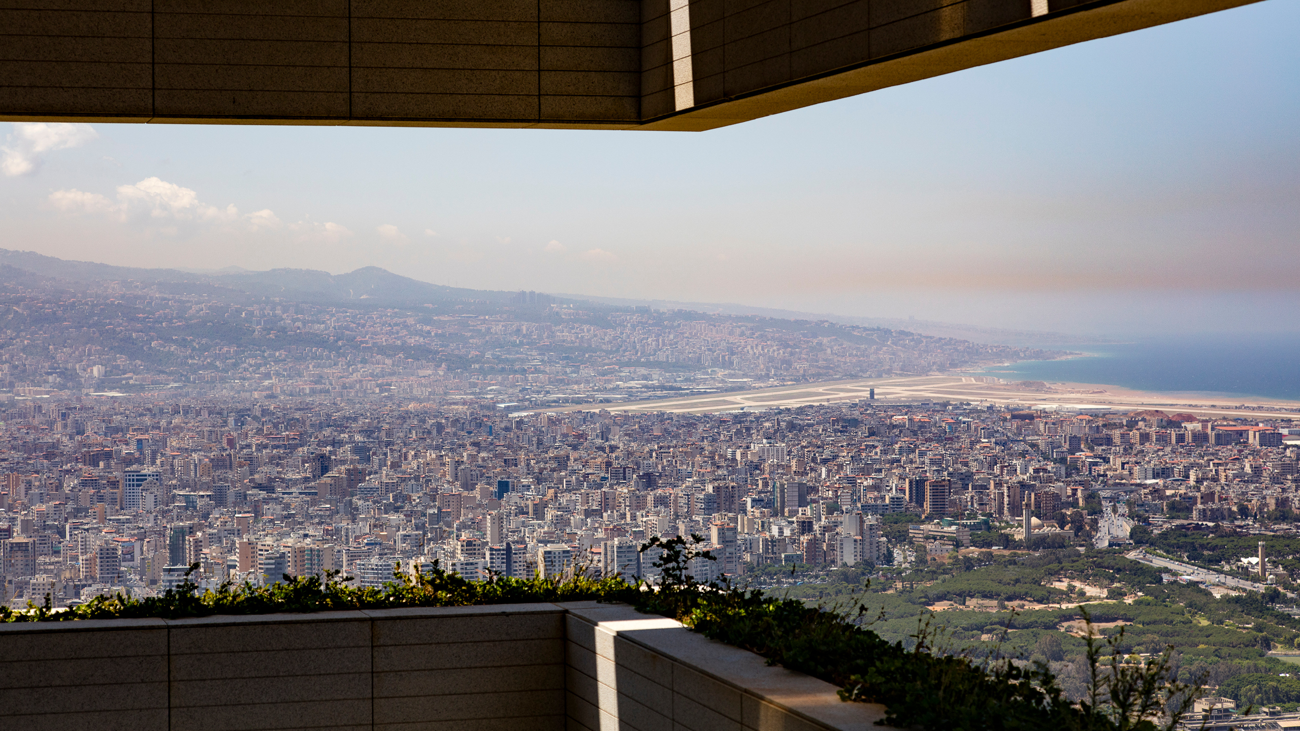Sweeping hilltop view from a Skygate terrace in Beirut over dense cityscape toward the coastline and airport runway in hazy daylight