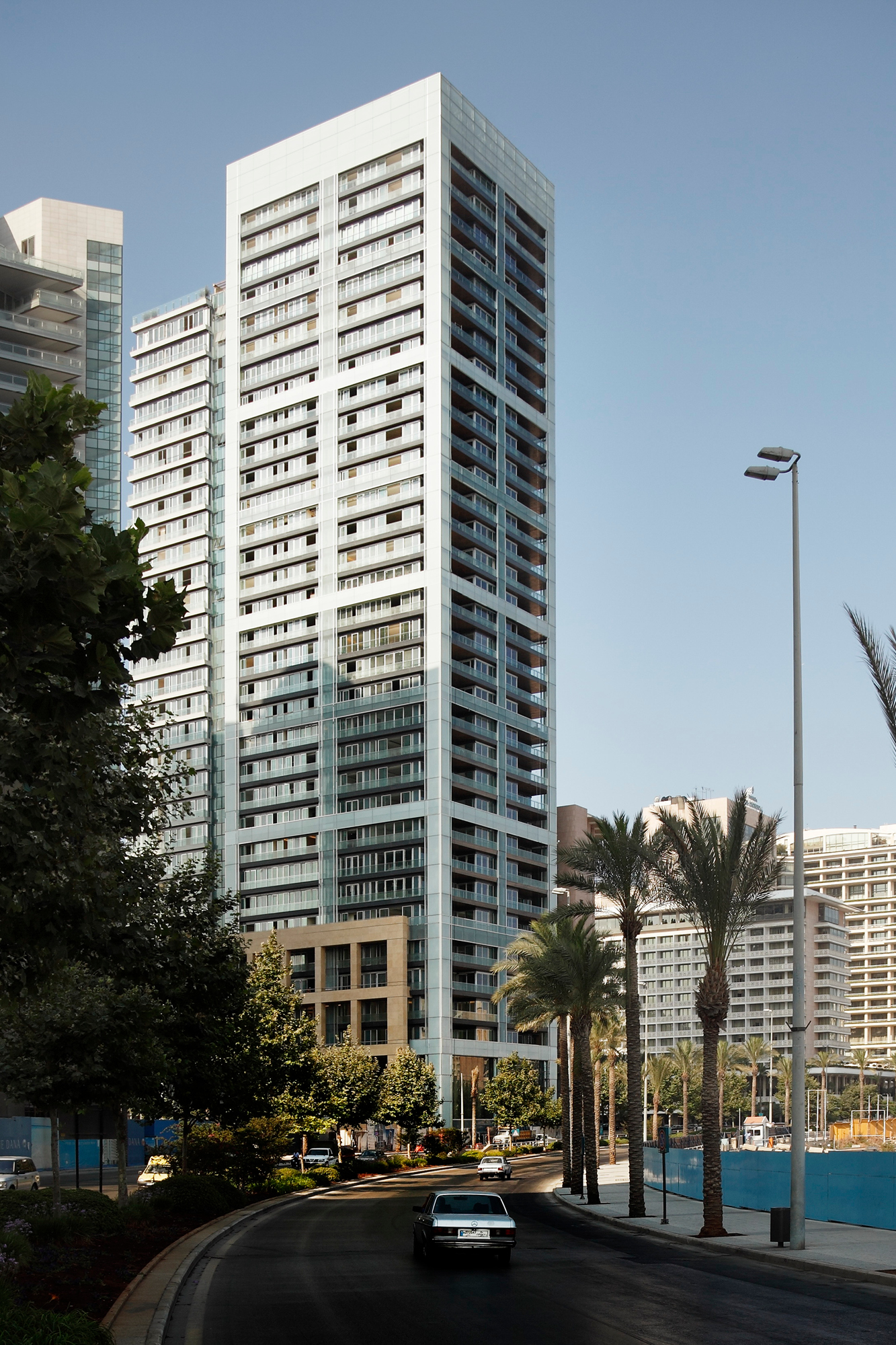 Platinum Tower in Beirut, a tall white high-rise with glass-cornered balconies, seen from a palm-lined street beside other coastal buildings