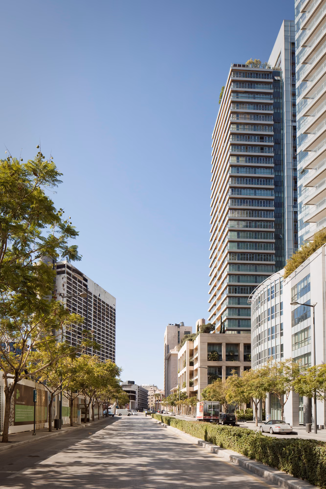 Platinum Tower rising above a tree-lined Beirut street, its glass-cornered balconies contrasting with lower stone buildings and a car-lined avenue