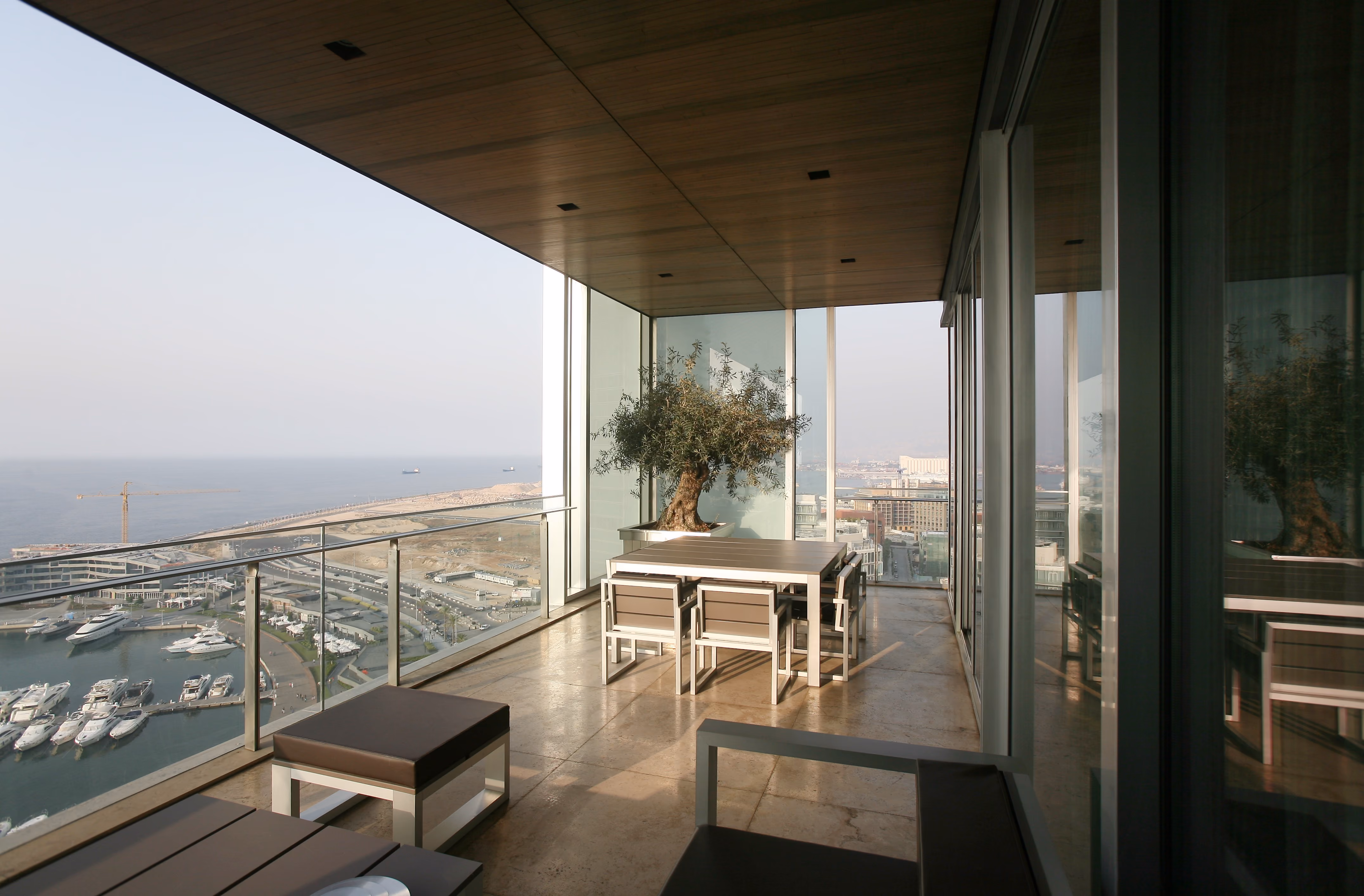 Wraparound balcony of Platinum Tower in Beirut, glass-enclosed terrace with dining set and potted olive tree overlooking the marina and coastline