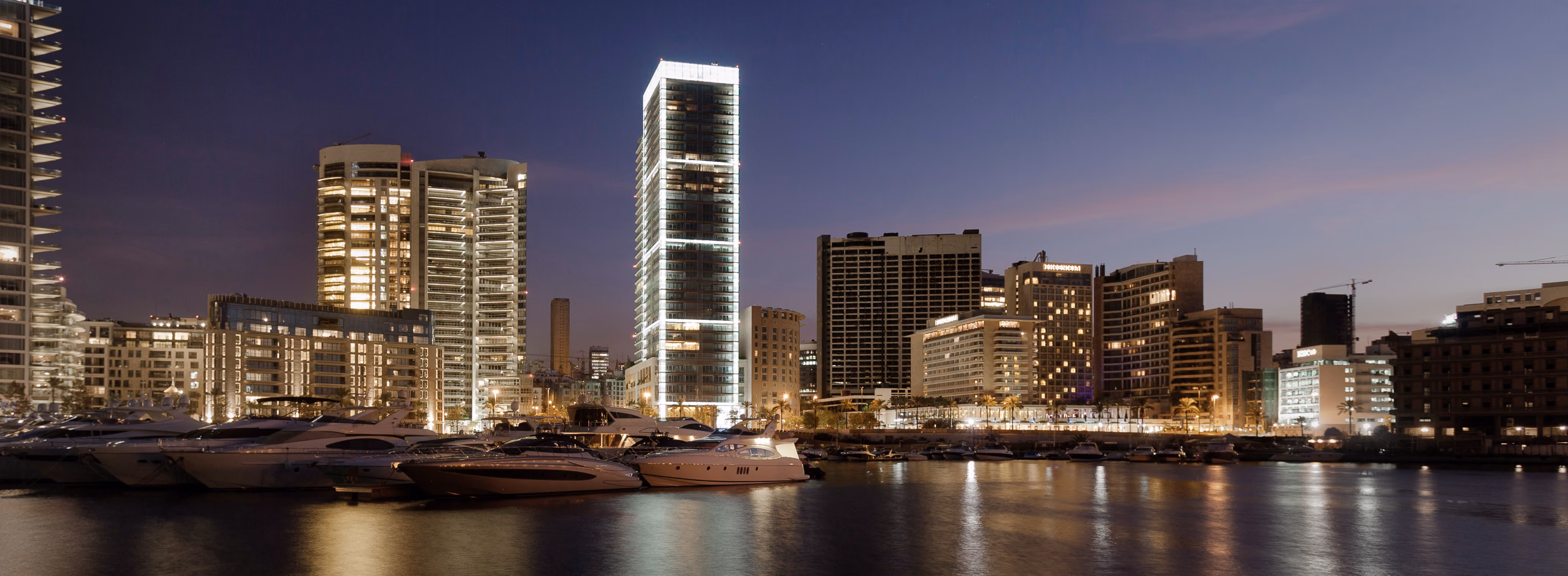 Platinum Tower in Beirut illuminated at dusk, its glass facade glowing above the marina skyline with yachts moored along the waterfront