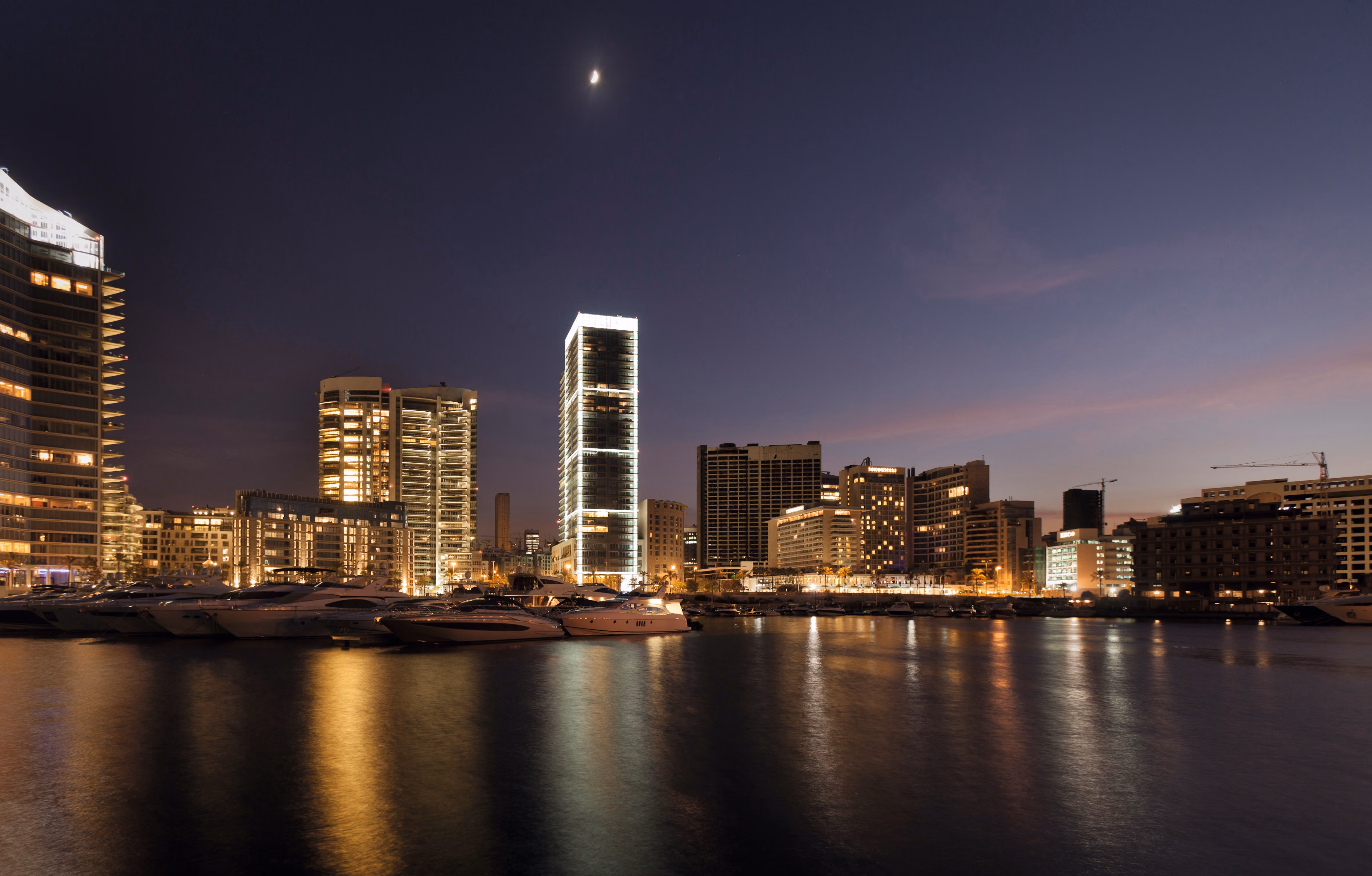 Platinum Tower in Beirut glowing white at dusk above the marina, surrounded by illuminated high-rises and yachts reflected in calm water