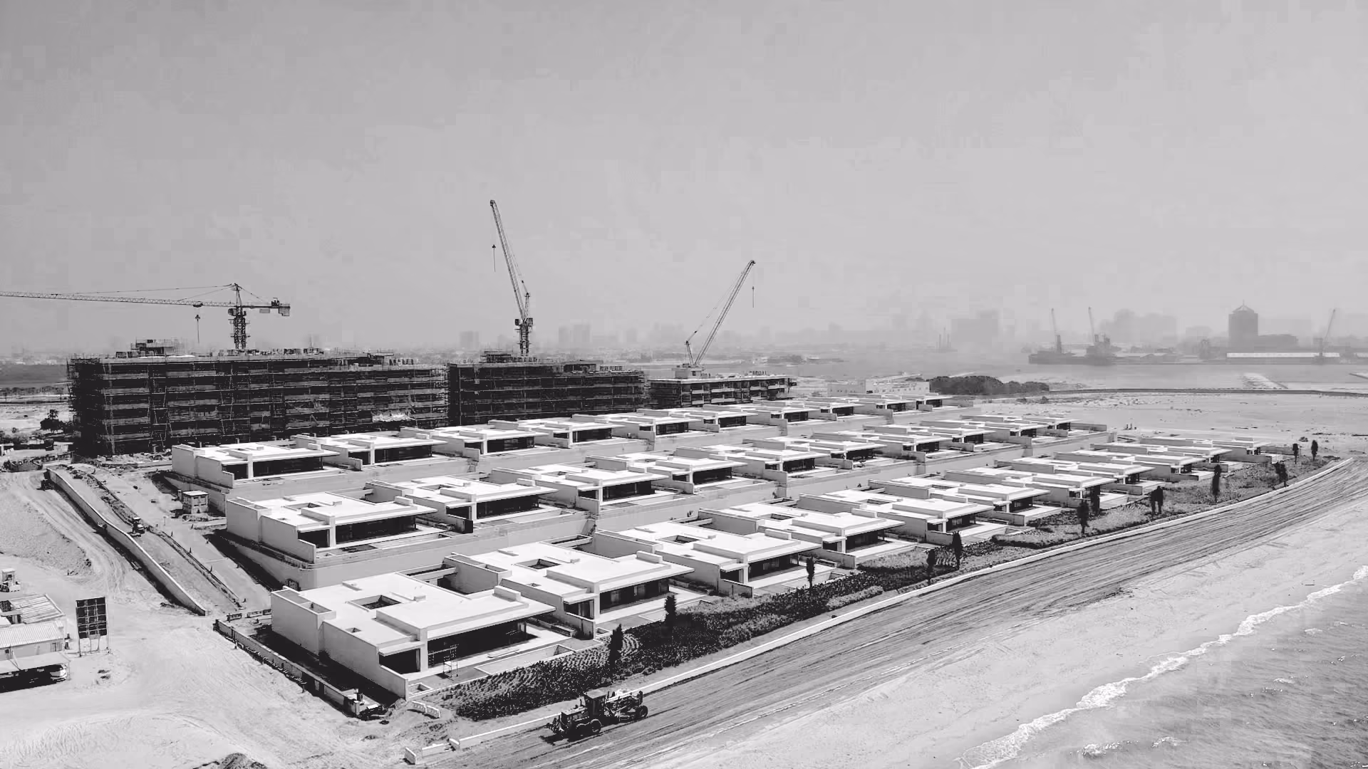 Black and white aerial view of Al Zorah Shores in Ajman under construction, rows of low-rise villas near the beach with cranes and mid-rise blocks beyond