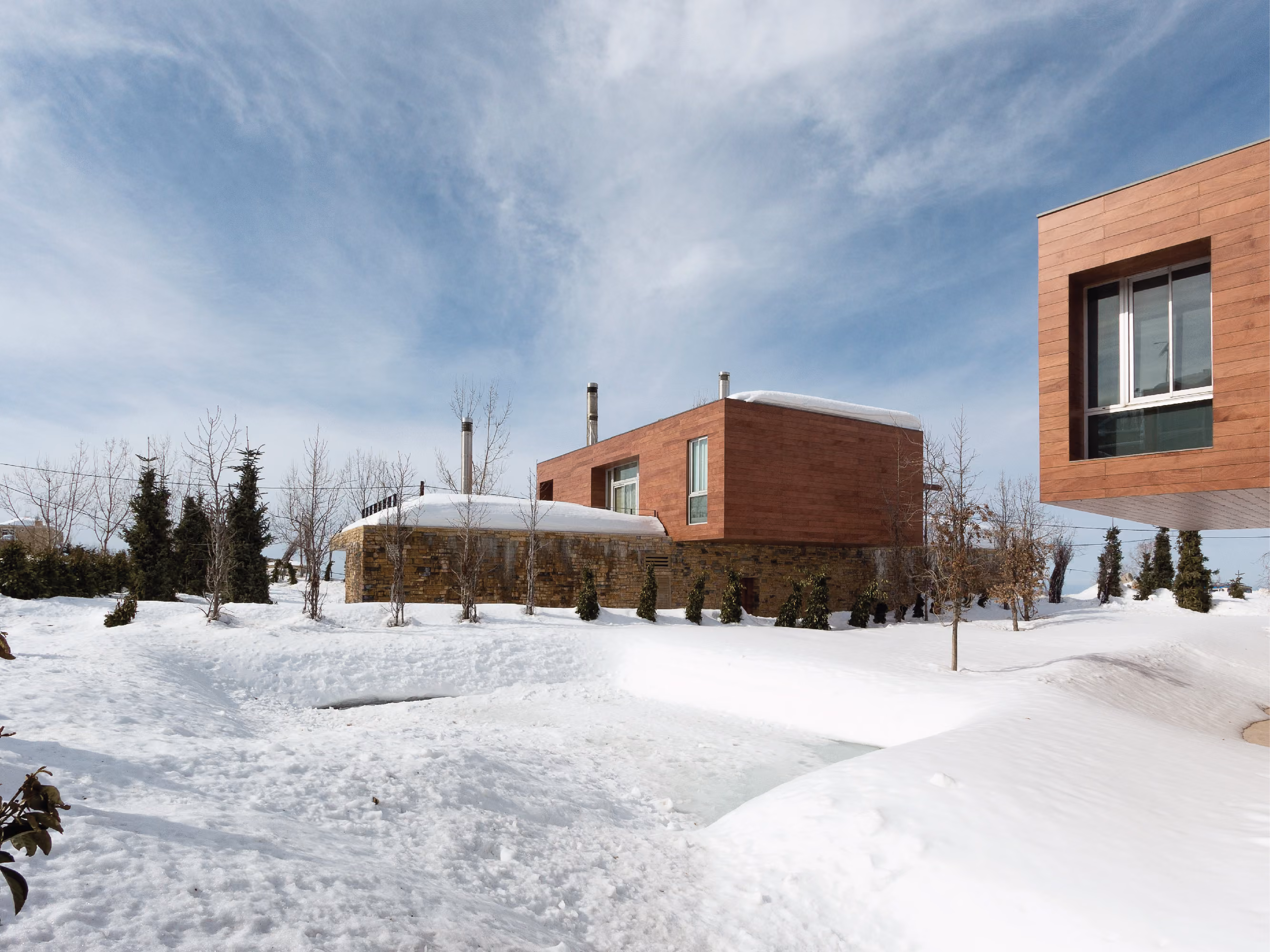 Snow-covered grounds at Clouds in Faqra, Lebanon, showing a wood and stone villa with cantilevered timber volume, sparse trees dotting the drifts