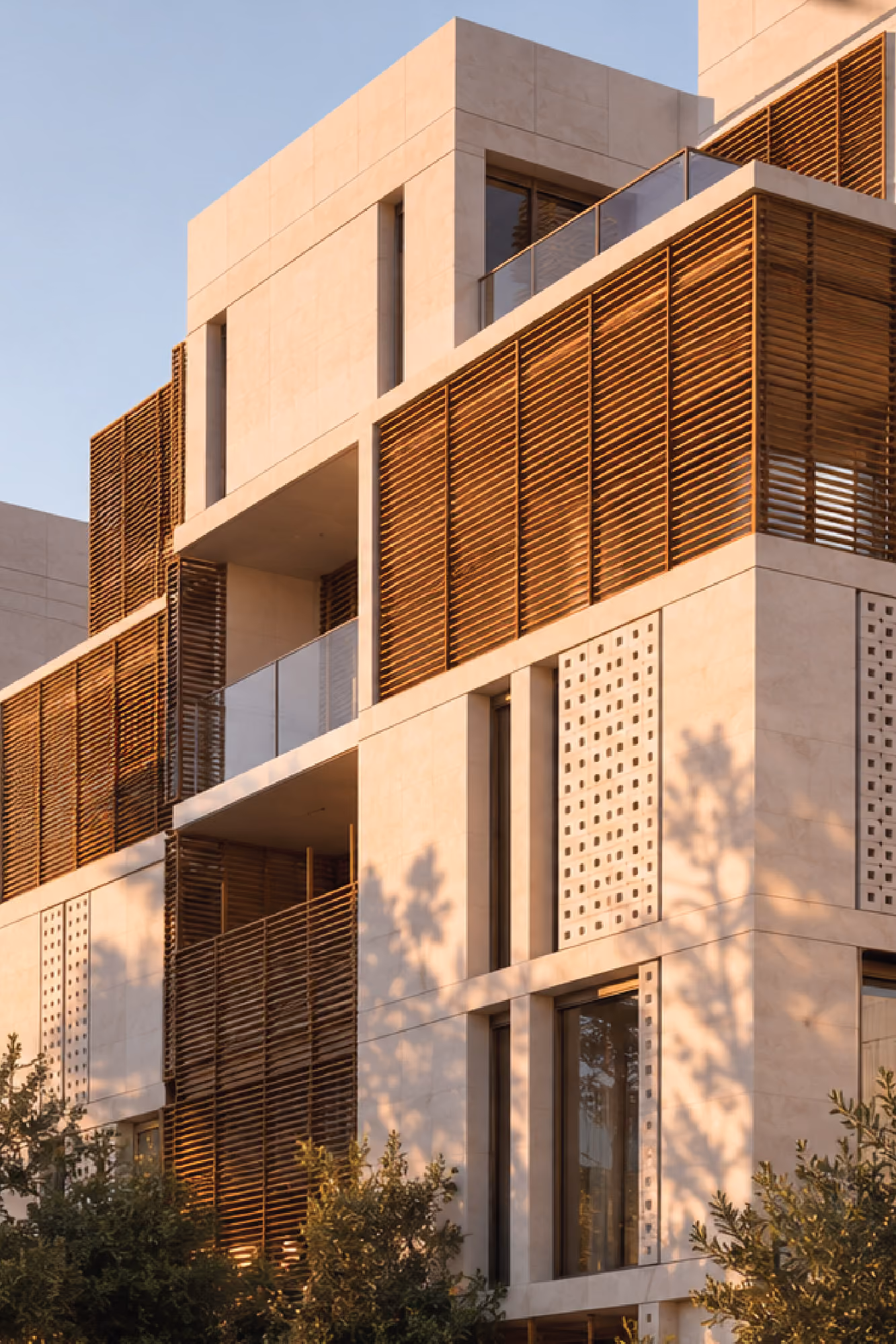 Facade detail of Doha Gardens in Al Khobar, stone-clad volumes with timber louvered screens and perforated metal panels catching warm evening light