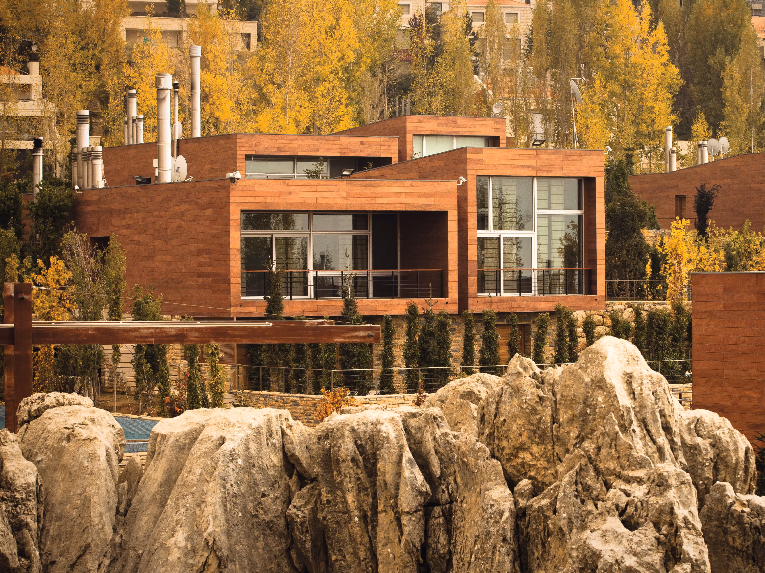 Cantilevered timber-clad villa at Clouds in Faqra, seen past large limestone boulders with autumn trees and hillside buildings behind