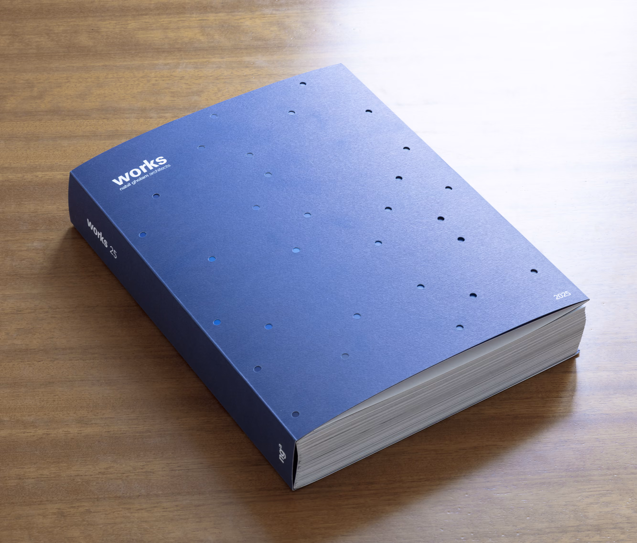 Cover of Works 25, the fifth Nabil Gholam Architects monograph, a thick blue hardback with scattered perforated dots resting on a wooden table
