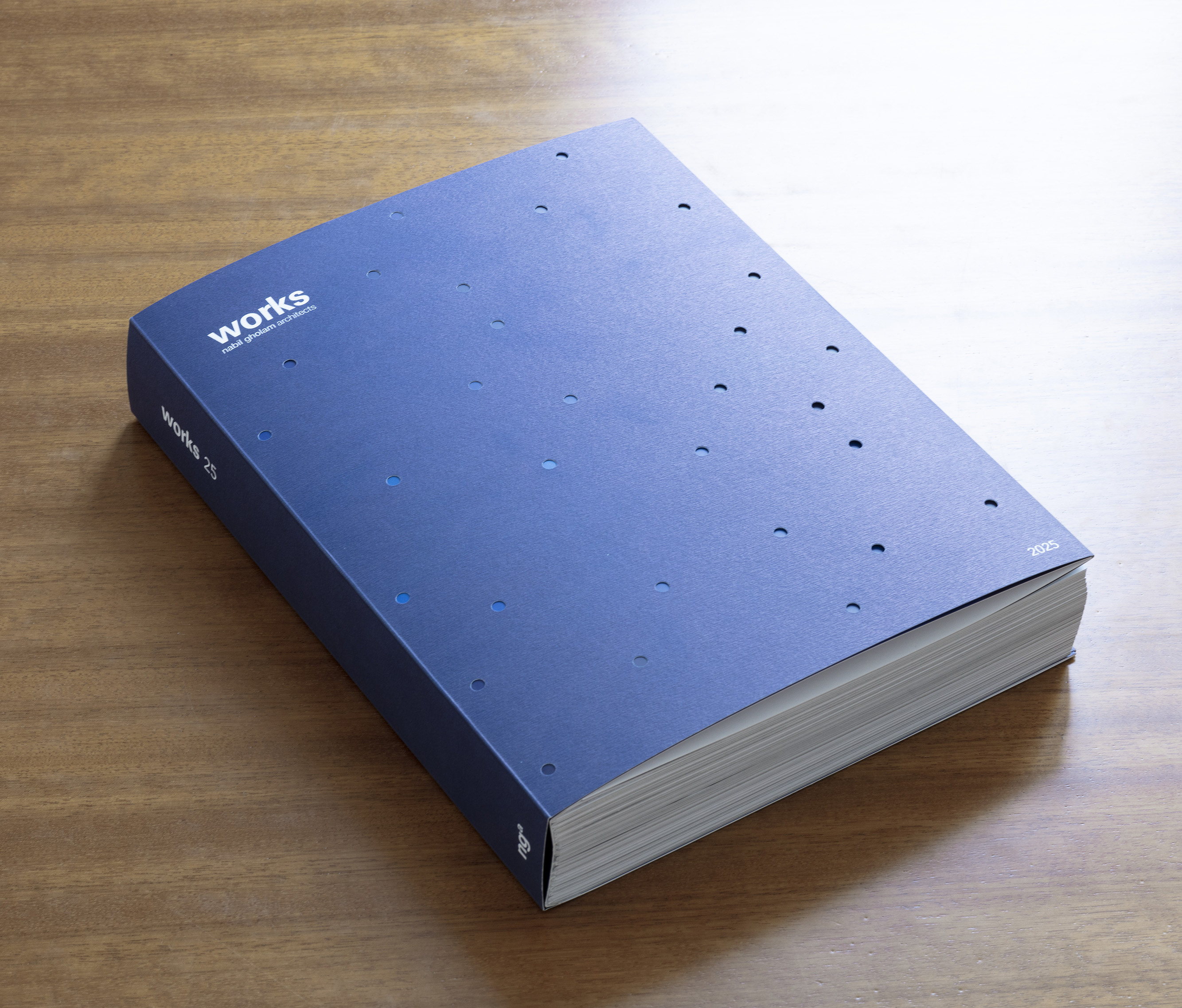Cover of Works 25, the fifth Nabil Gholam Architects monograph, a thick blue hardback with scattered perforated dots resting on a wooden table