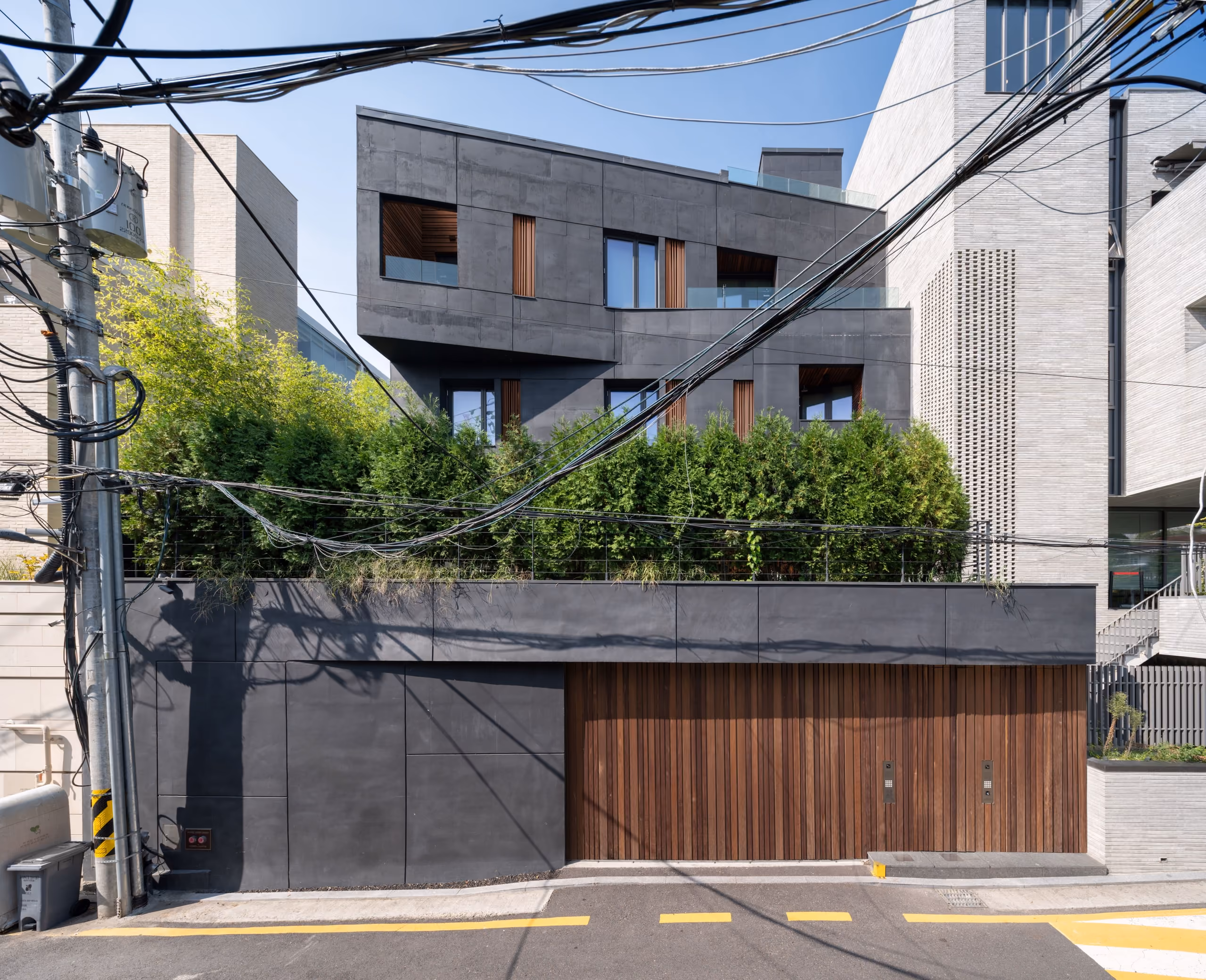 Dark faceted concrete facade of Pyrite, a private residence in Seoul, with timber-slatted garage doors and greenery screening the street-level wall