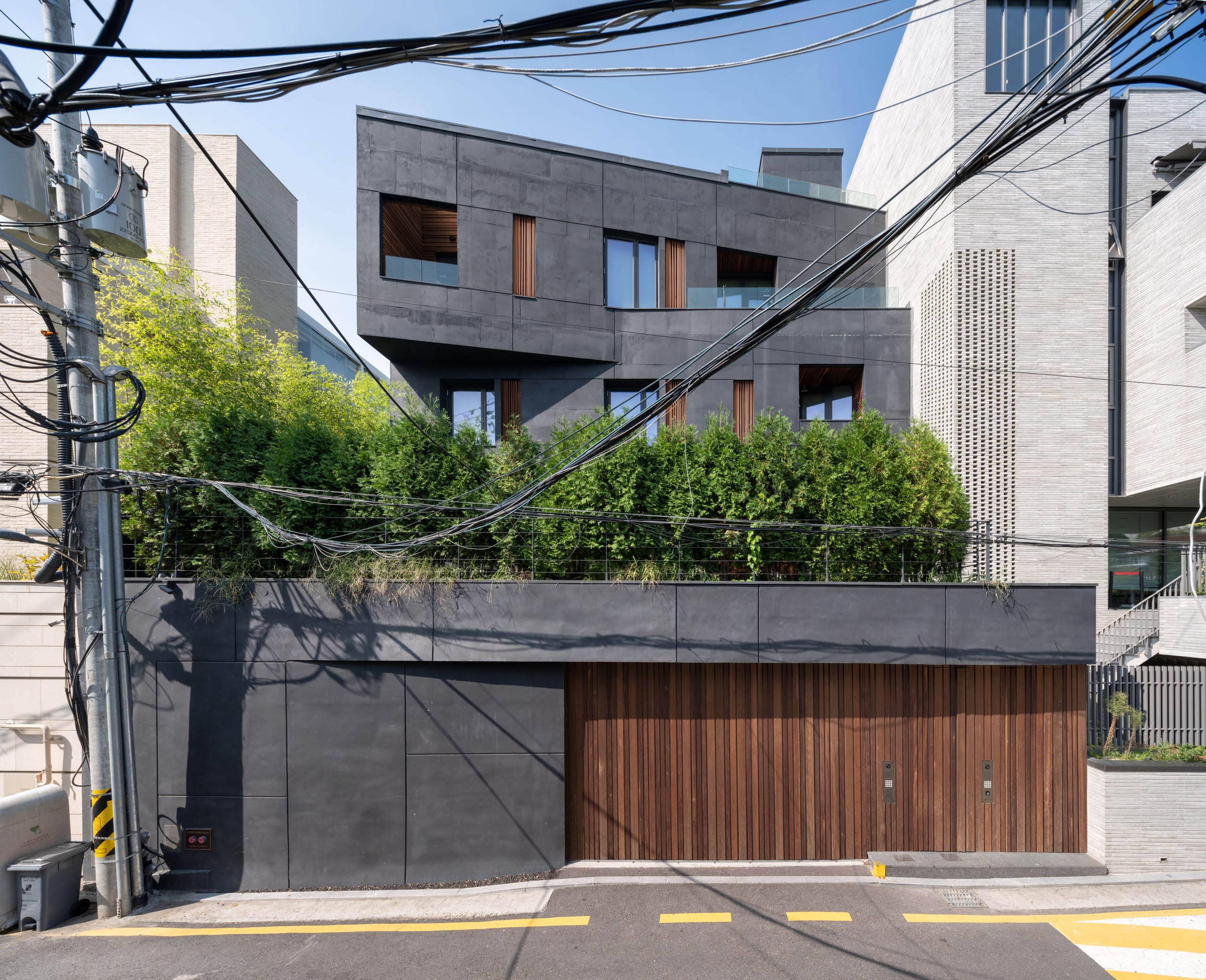 Dark faceted concrete facade of Pyrite, a private residence in Seoul, with timber-slatted garage doors and greenery screening the street-level wall