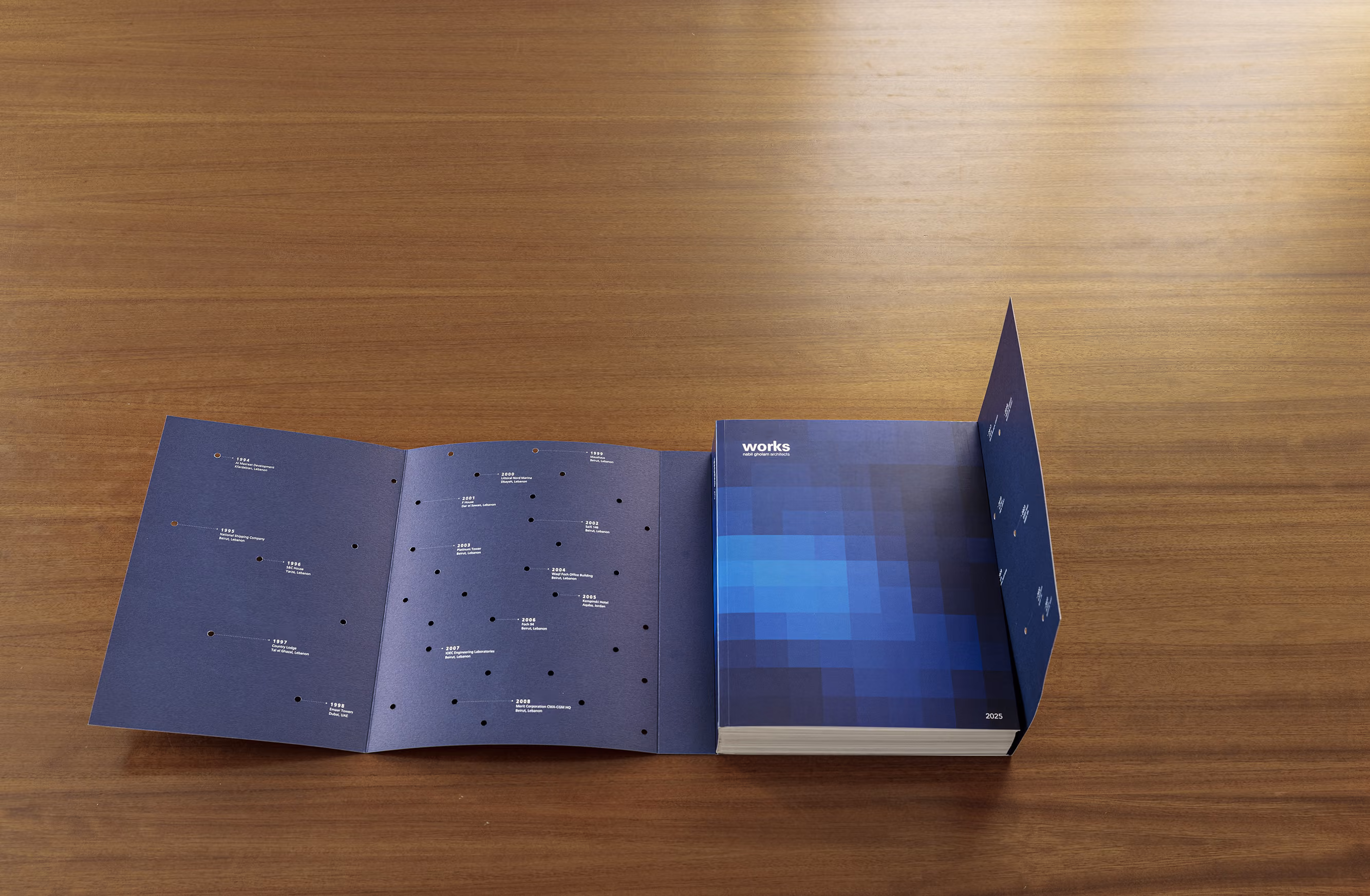 Works 2025 monograph on a wooden table, blue pixelated cover beside an unfolded timeline page with dated project milestones