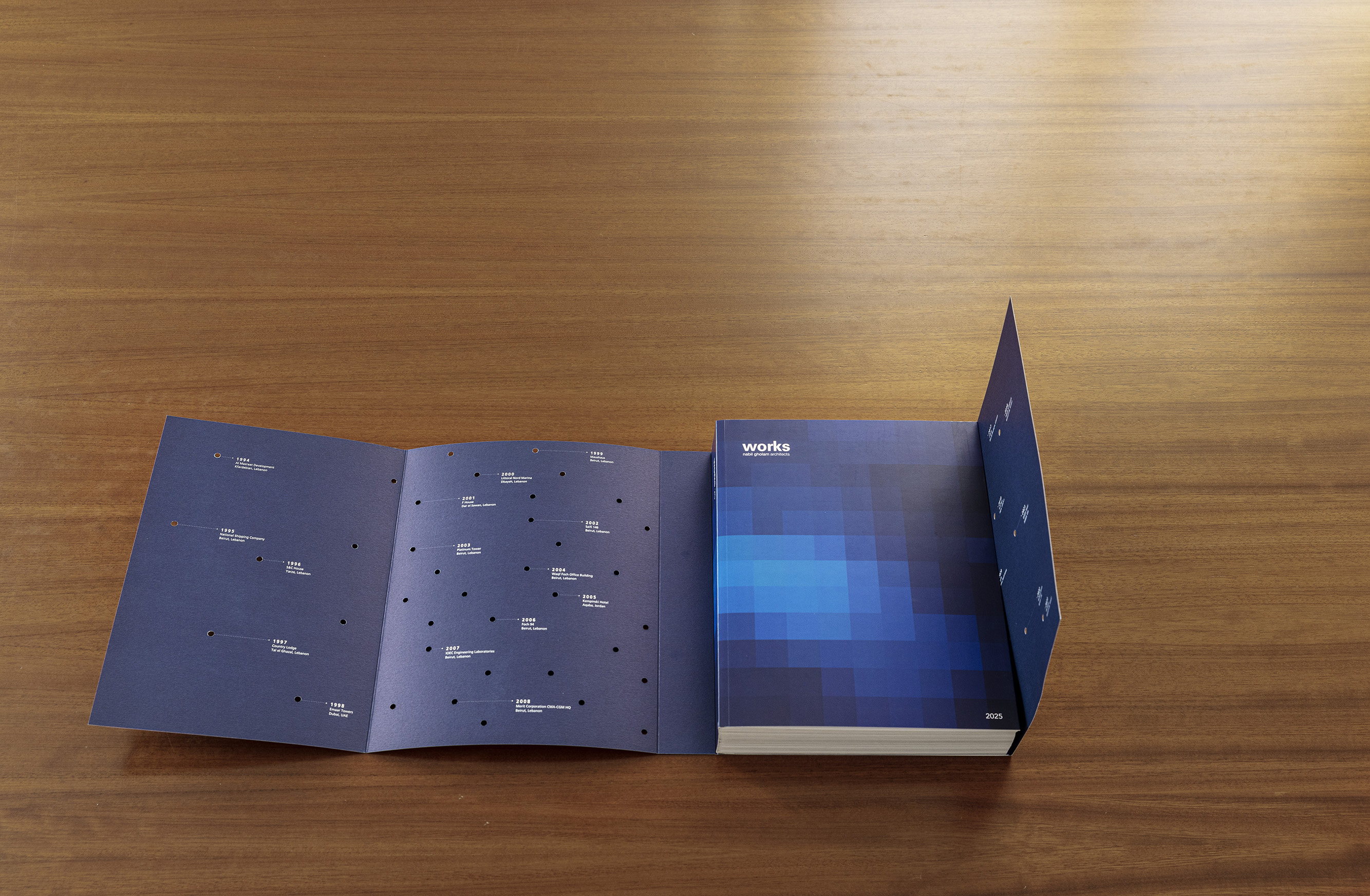 Works 2025 monograph on a wooden table, blue pixelated cover beside an unfolded timeline page with dated project milestones
