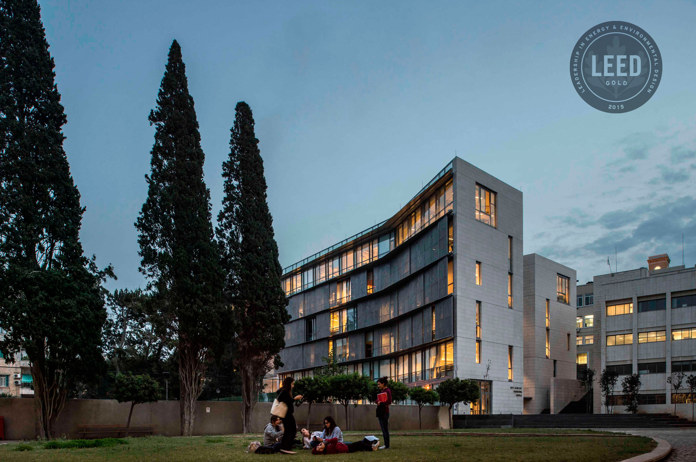 Curved stone and glass academic building lit at dusk, with cypress trees and students gathered on the lawn, LEED Gold certification badge overlaid