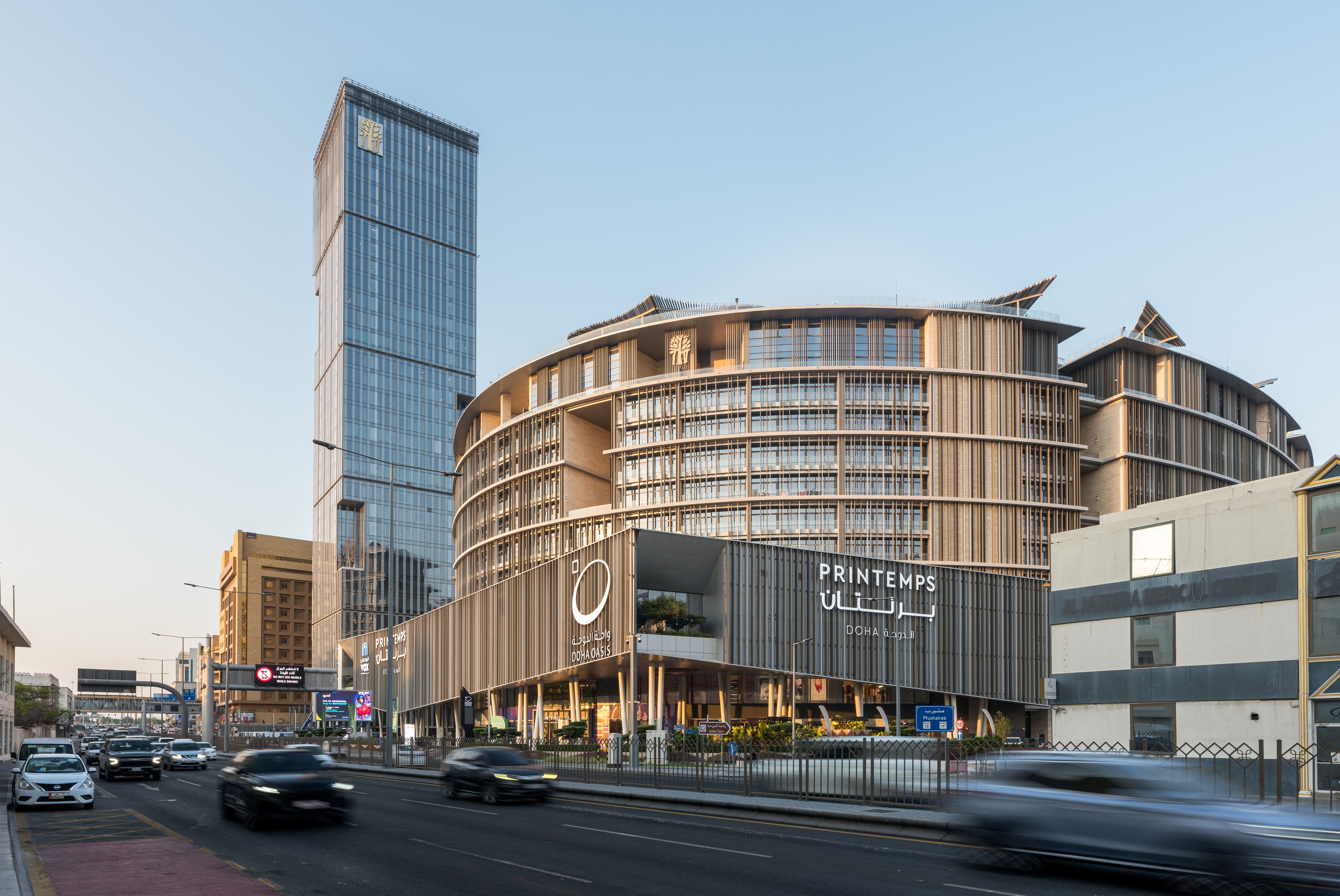 Curved timber-slatted building housing Doha Oasis and Printemps, with a slender glass high-rise behind, part of a mixed-use development by Nabil Gholam Architects