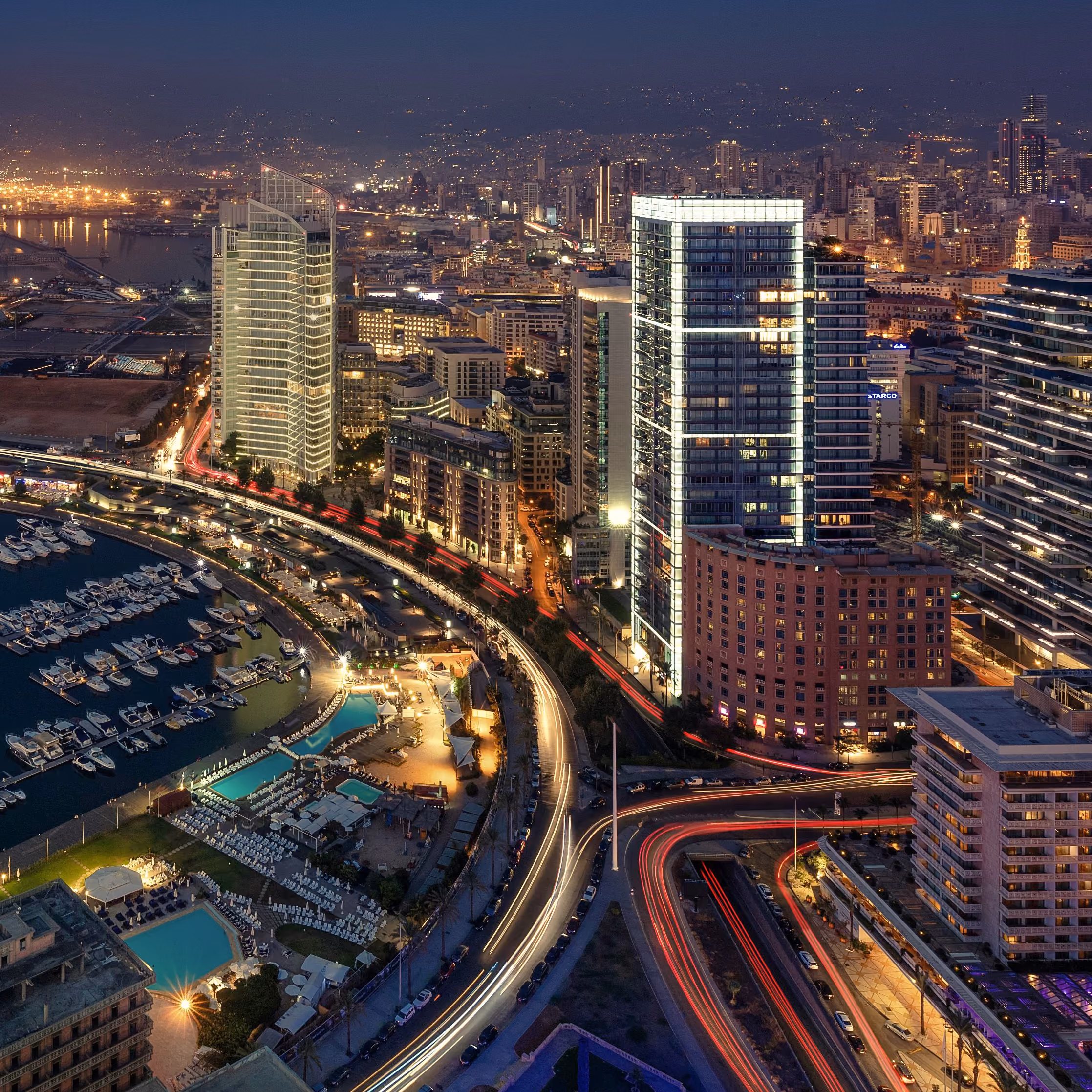 Aerial night view of Beirut's high-rise skyline along the coastal corniche, illuminated towers, marina and looping highways with light trails from traffic