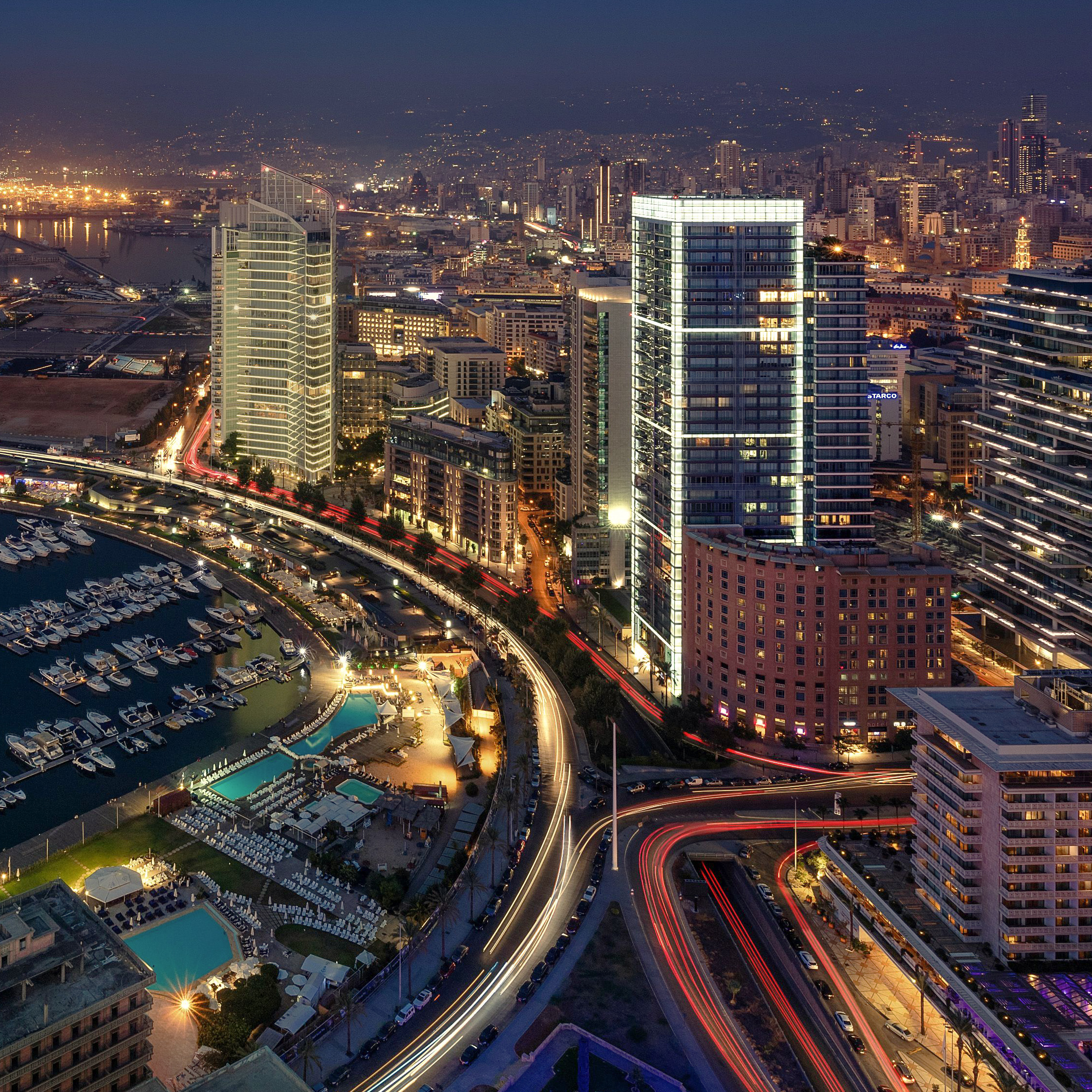 Aerial night view of Beirut's high-rise skyline along the coastal corniche, illuminated towers, marina and looping highways with light trails from traffic