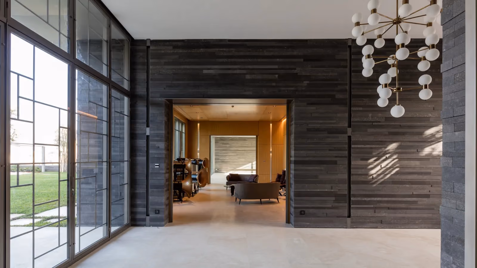 AY House interior design in Yarze, Lebanon, by Nabil Gholam Architects, layered basalt-clad walls, floor-to-ceiling glazing and a sculptural globe chandelier leading to a warm lounge beyond