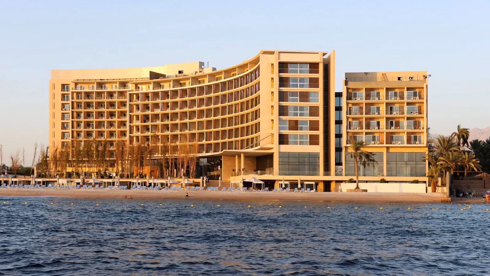 Kempinski Hotel by Nabil Gholam Architects, a curved beachfront hotel in golden stone rising along the shoreline in Aqaba, Jordan