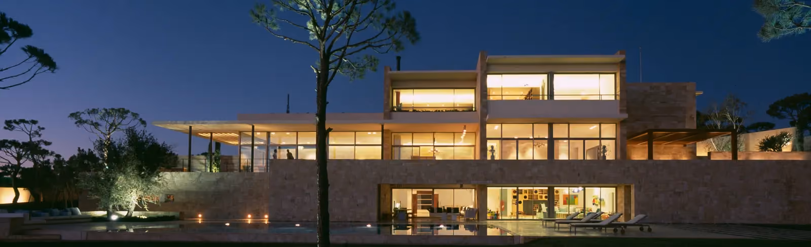 F House by Nabil Gholam Architects, illuminated at dusk in Dahr el Sawan, Lebanon, stone walls and glazed volumes overlooking a reflecting pool and pine trees