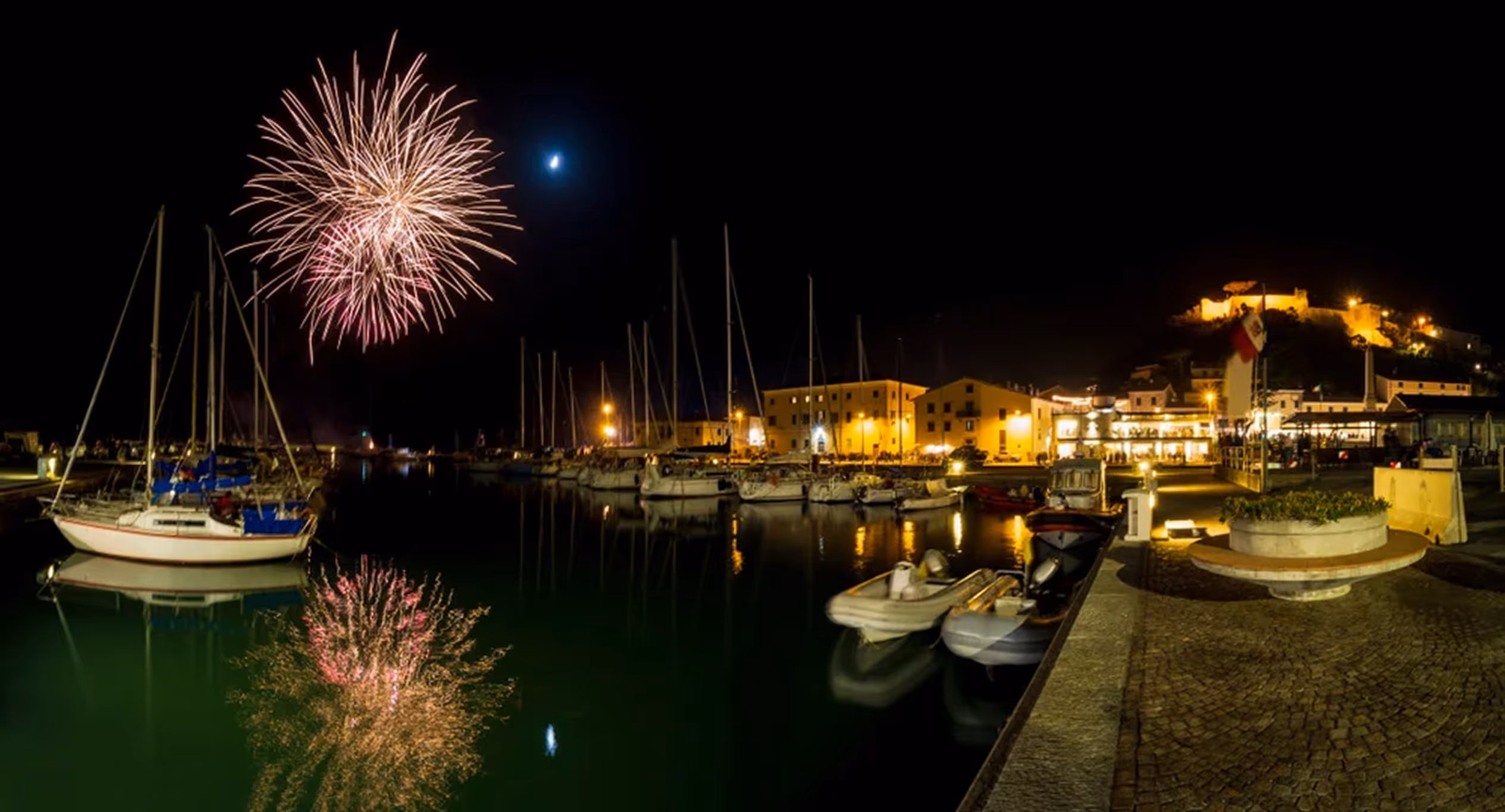 Святковий феєрверк над яхтовою пристанню з відображенням у воді і освітленими будівлями вночі.