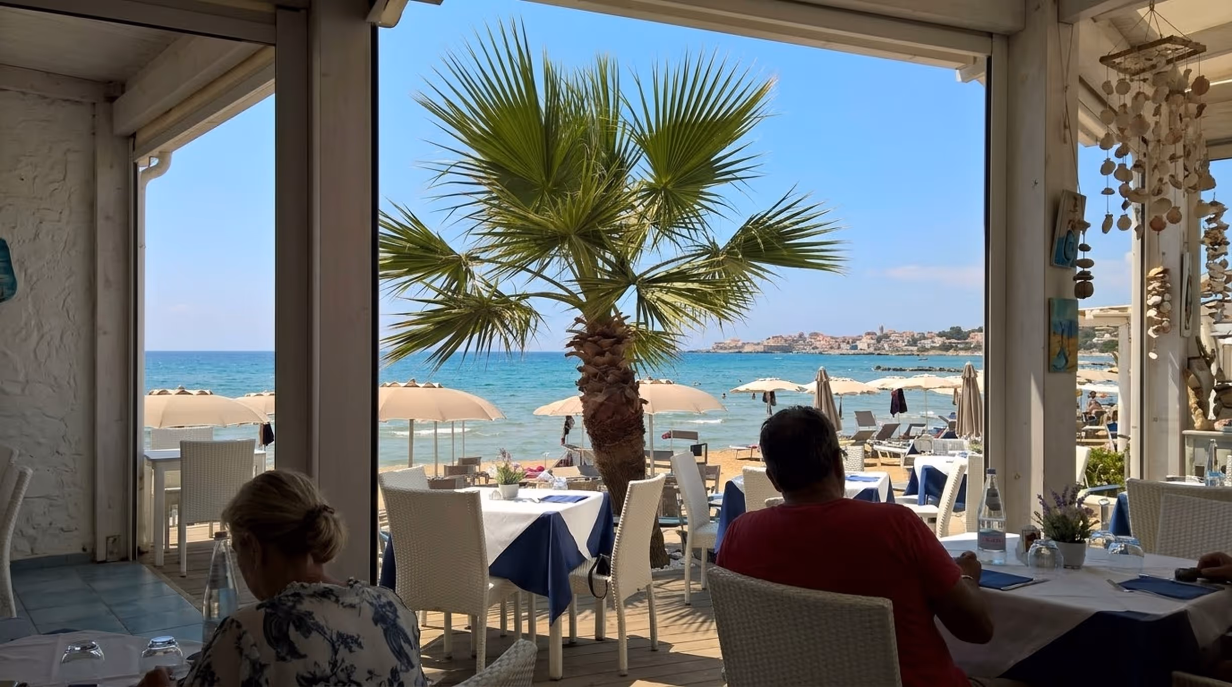 Вид на пляж з парасольками та морем з відкритої тераси ресторану з білим плетеним меблями й столами, накритими біло-синіми скатертинами.