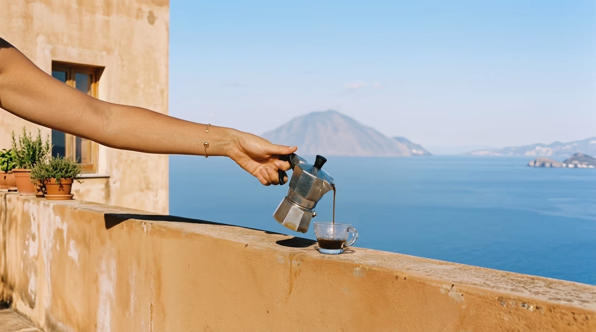 Рука наливає каву з металевого кавника в скляну чашку на терасі з видом на море та гору на горизонті.