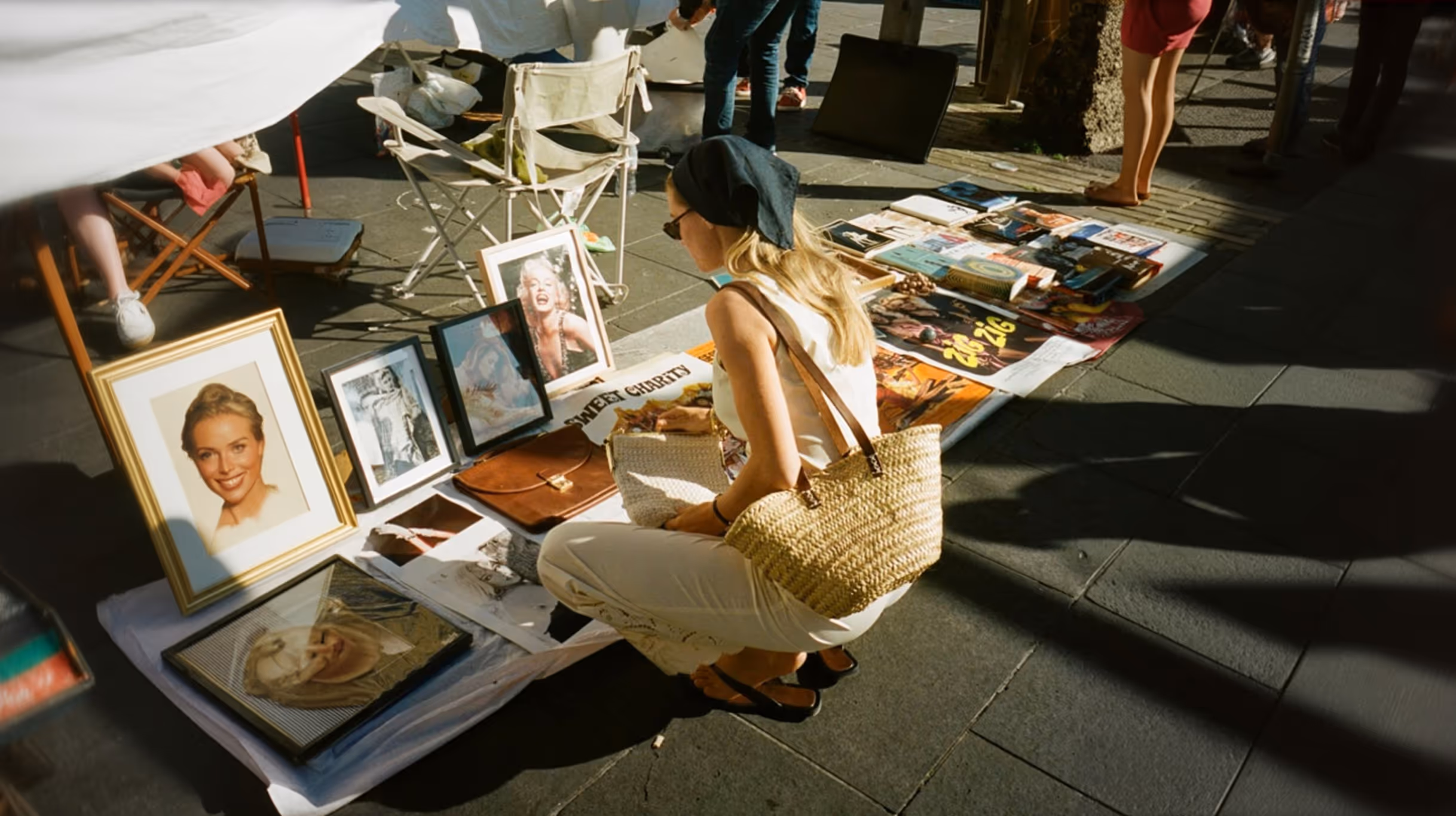 Жінка в світлому одязі і чорній хустці присіла оглянути картини та предмети, виставлені на вуличному базарі на тротуарі.