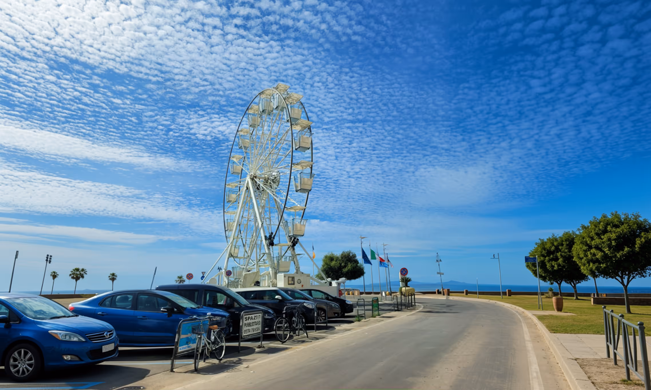 Колесо огляду біля набережної з припаркованими автомобілями під блакитним небом із хмарками.