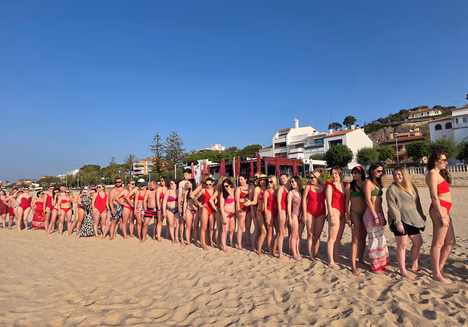 Велика група людей у купальниках стоїть у чергу на піщаному пляжі на фоні будинків під ясним блакитним небом. Enjoy 2026, Інтенсив MIGIS, інтенсив для гештальт-терапевтів