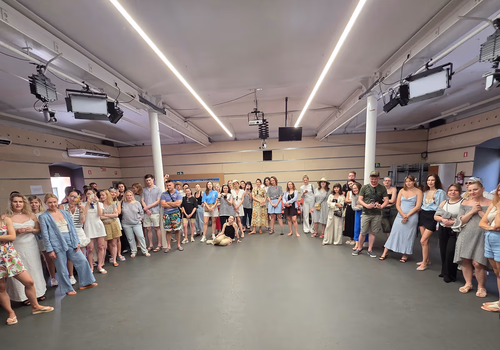 Велика група людей стоїть півколом в просторій кімнаті з бежевими стінами та світлодіодними панелями на стелі. Enjoy 2026, Інтенсив MIGIS, інтенсив для гештальт-терапевтів