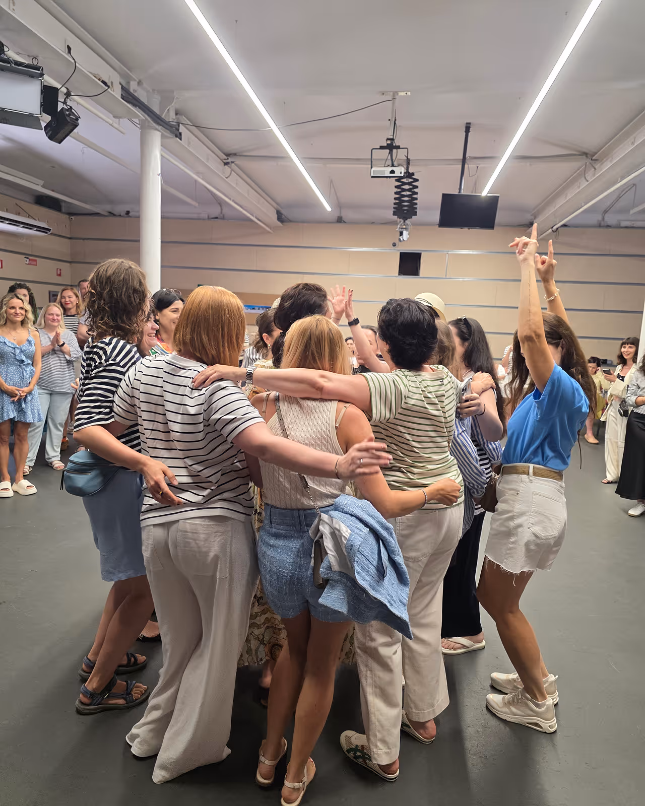 Група жінок обіймається та радіє у просторій кімнаті з іншими людьми на тлі. Enjoy 2026, Інтенсив MIGIS, інтенсив для гештальт-терапевтів