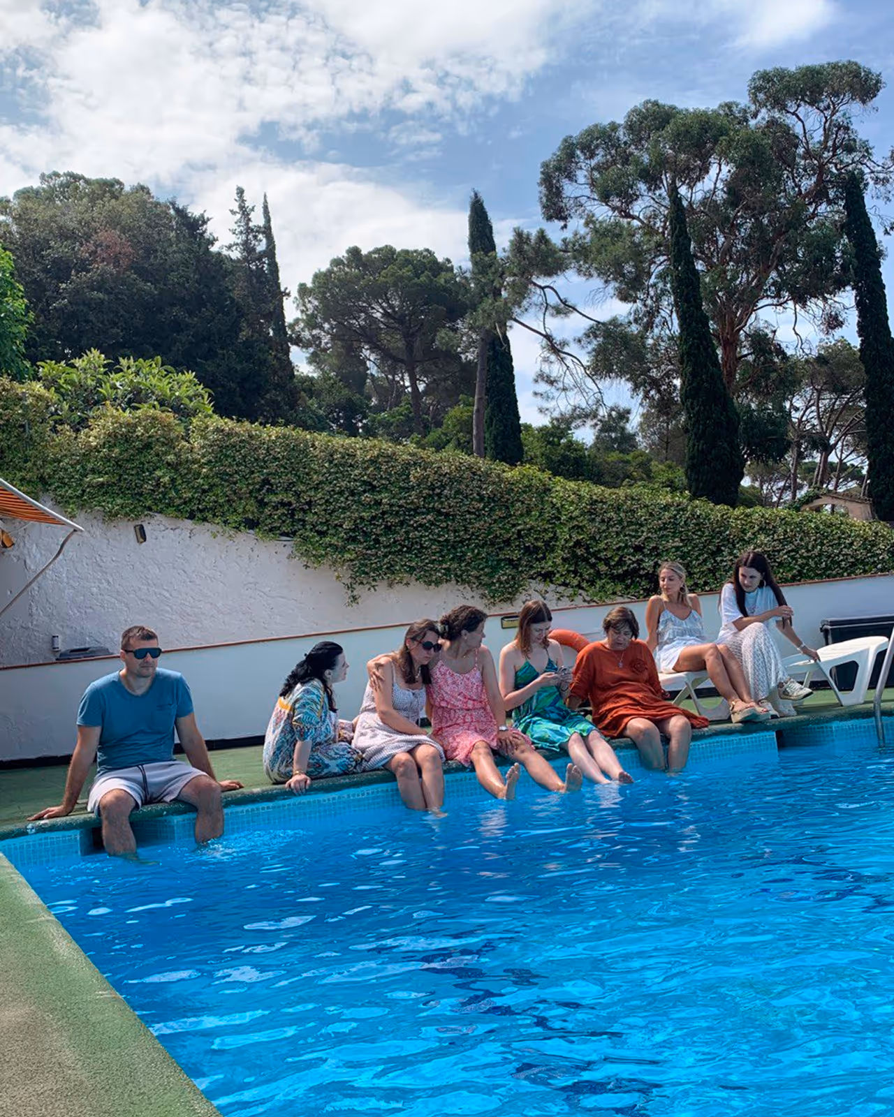 Група людей сидить біля краю басейну, занурюючи ноги у воду на тлі зелені та блакитного неба. Enjoy 2026, Інтенсив MIGIS, інтенсив для гештальт-терапевтів