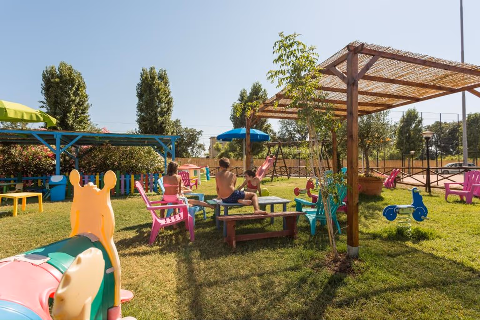 Діти граються біля столу з кольоровими кріслами на відкритому дитячому майданчику з гойдалками, гіркою та ігровими конструкціями на траві під сонячним небом. Villaggio Mare Si