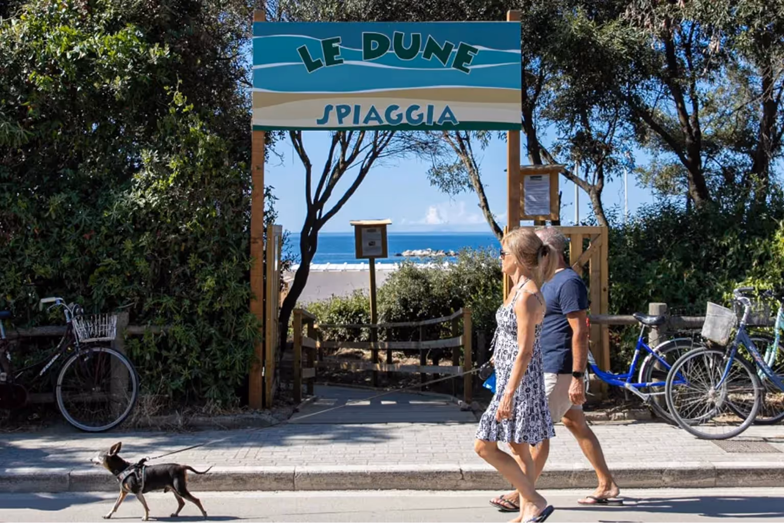 Пара іде з маленьким собакою на повідку повз вхід до пляжу Le Dune з видом на море та велосипедами з боків. Villaggio Mare Si