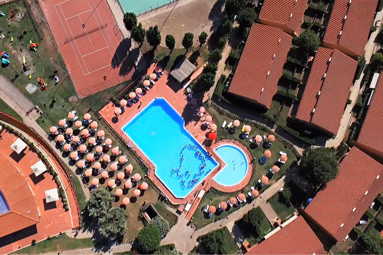 Вид зверху на басейн із сонячними парасолями навколо та сусідній тенісний корт в озелененій зоні з будинками з червоними дахами. Villaggio Mare Si