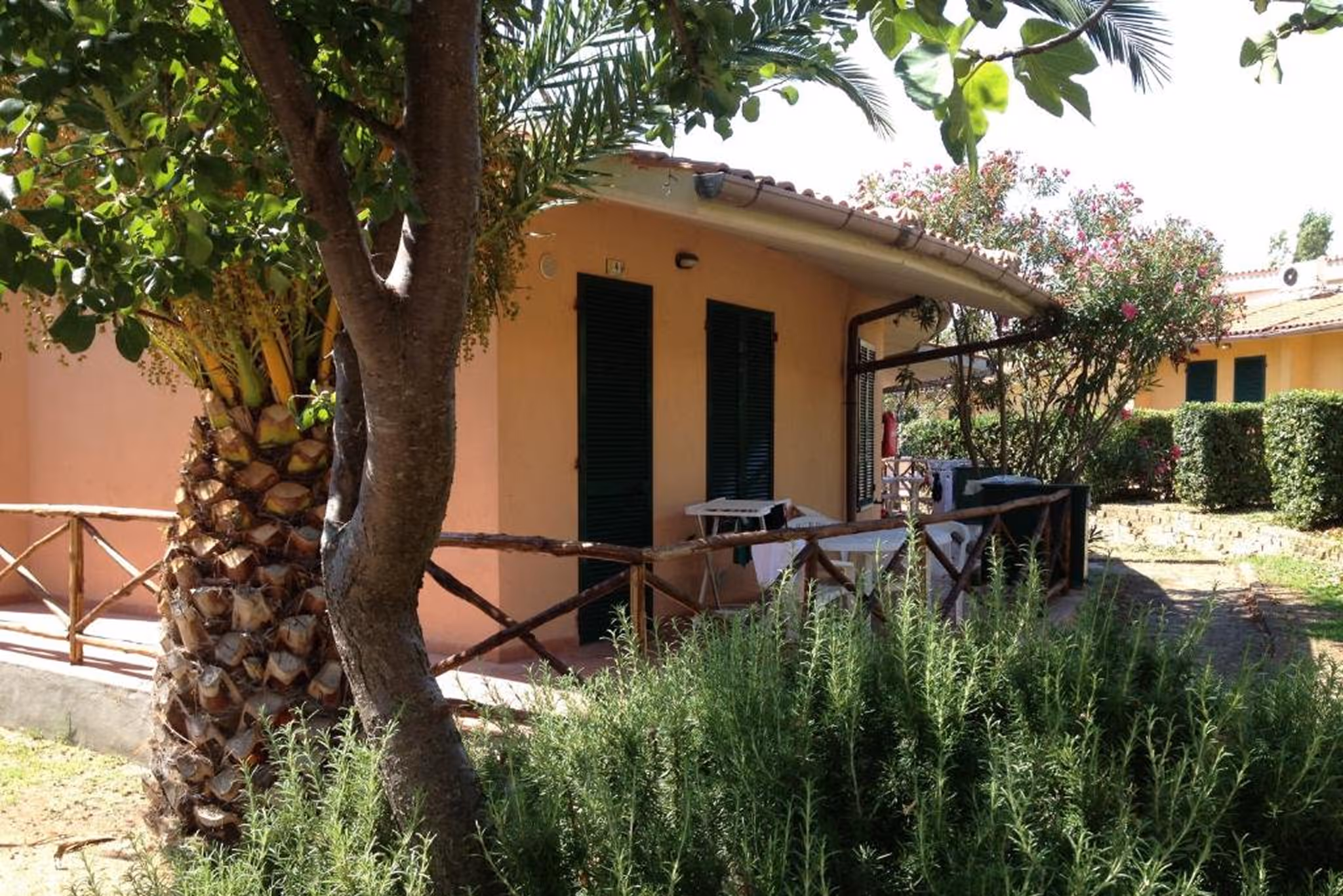Тераса помаранчевого будинку з закритими темно-зеленими ставнями та дерев'яним огородженням посеред саду з пальмами і кущами. Villaggio Mare Si
