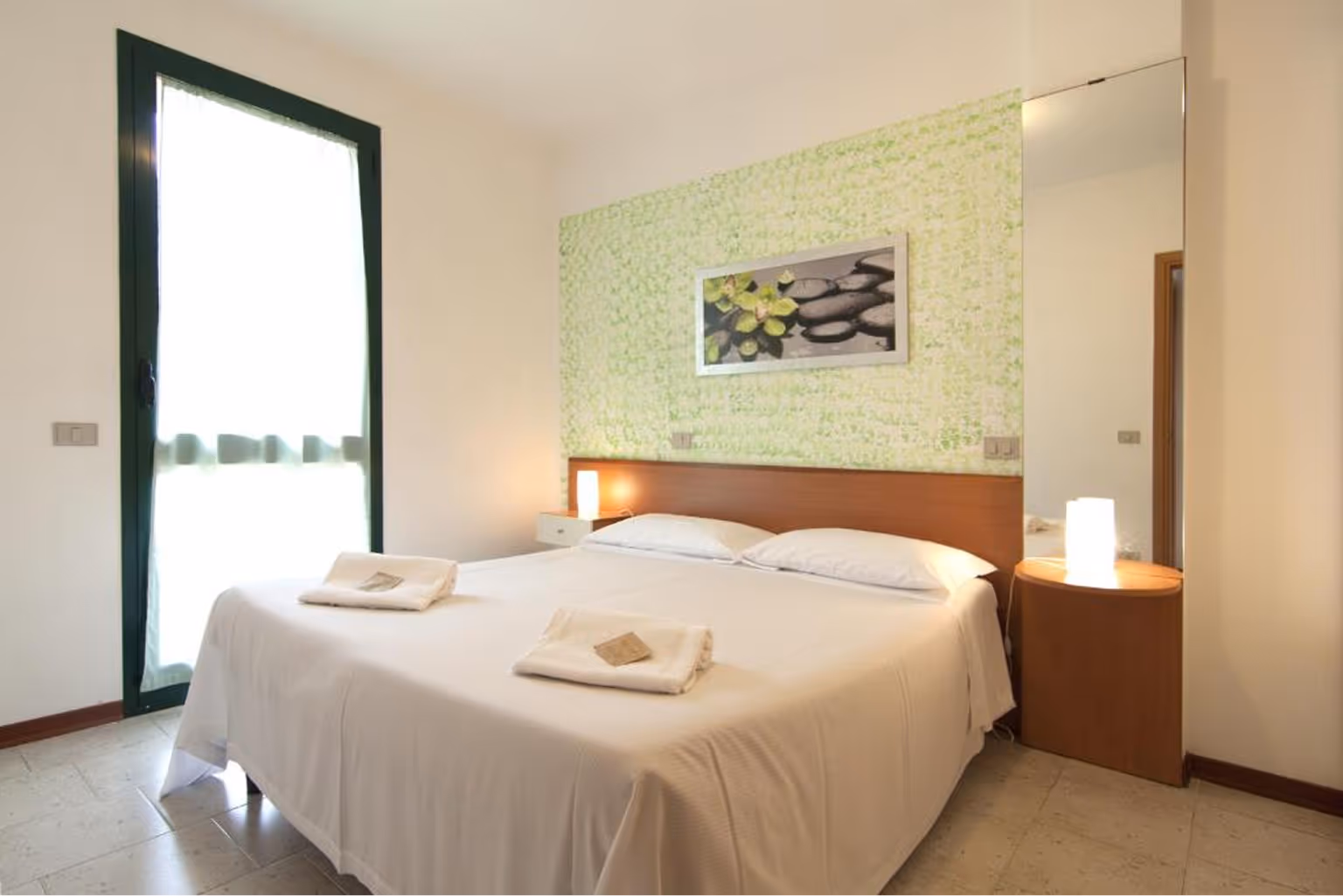 Світла спальня з двоспальним ліжком, білою постільною білизною, двома складеними рушниками та нічними лампами біля ліжка. Villaggio Mare Si